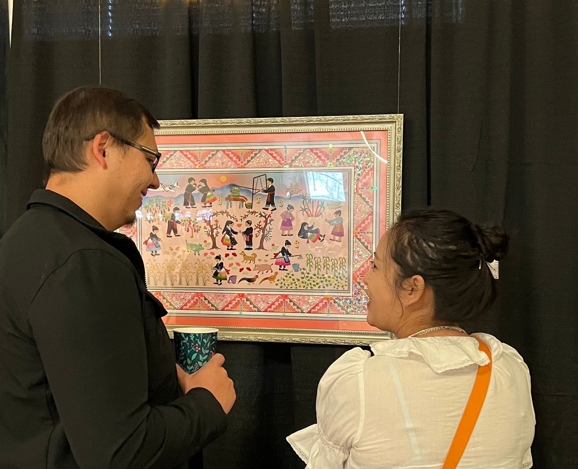Two people looking at a framed colorful painting in an art exhibit. Man holds a cup, woman smiles.