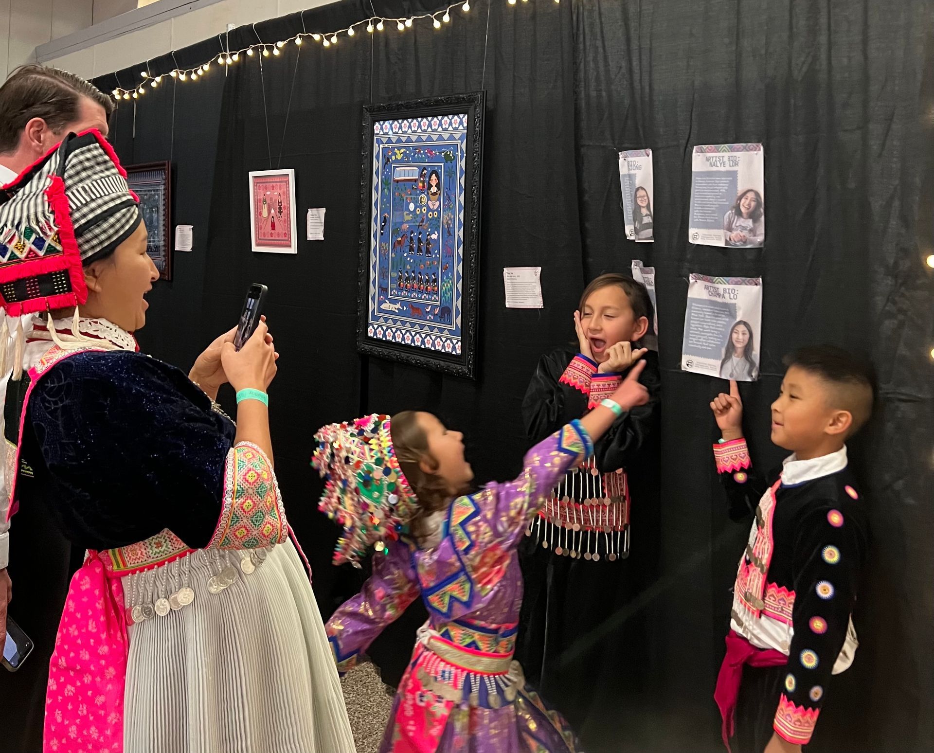 People in colorful traditional clothing taking photos in front of artwork displayed on a black wall.