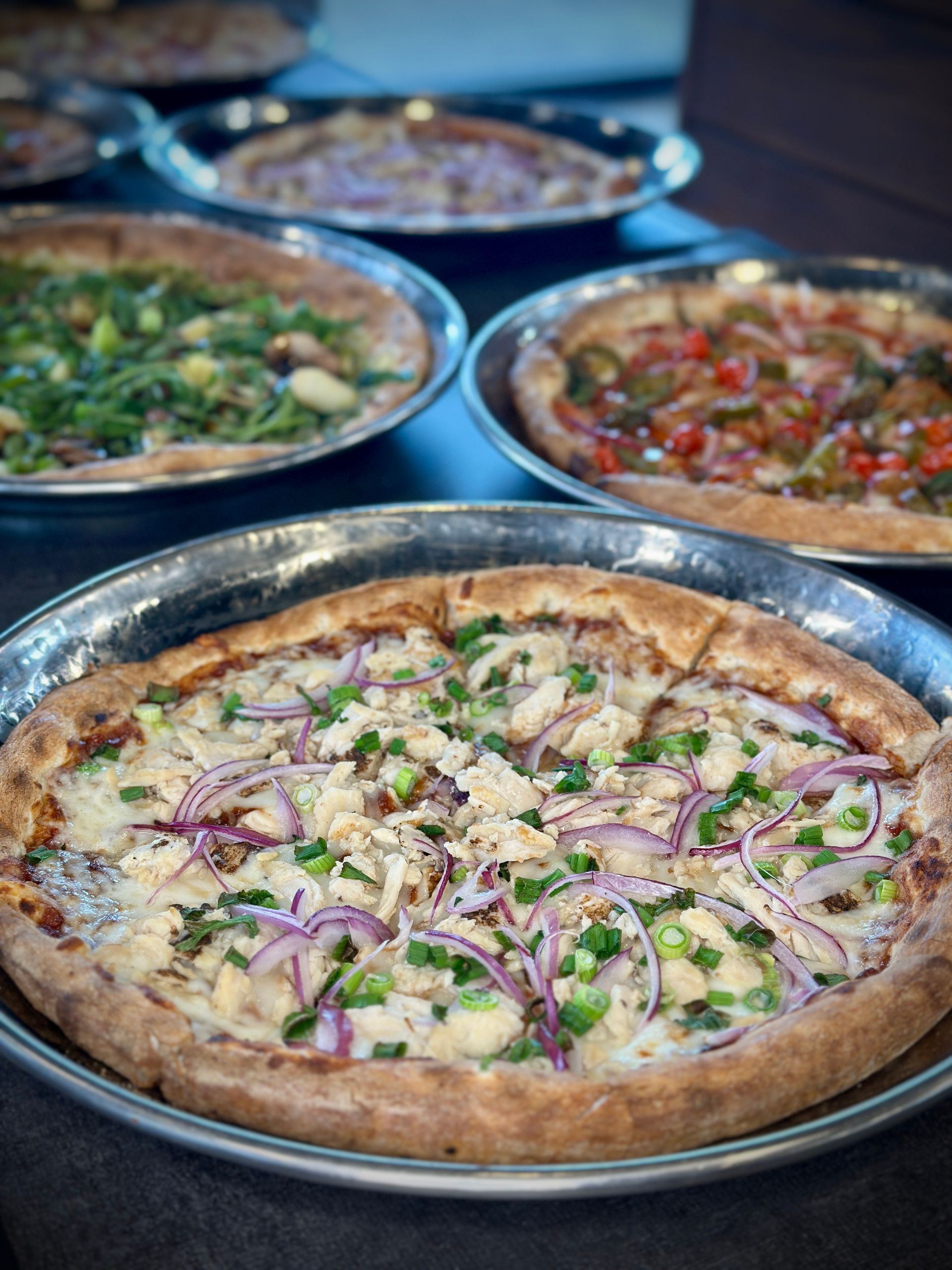 Several pizzas topped with ingredients like chicken, red onions, and peppers on silver trays set on a dark surface.