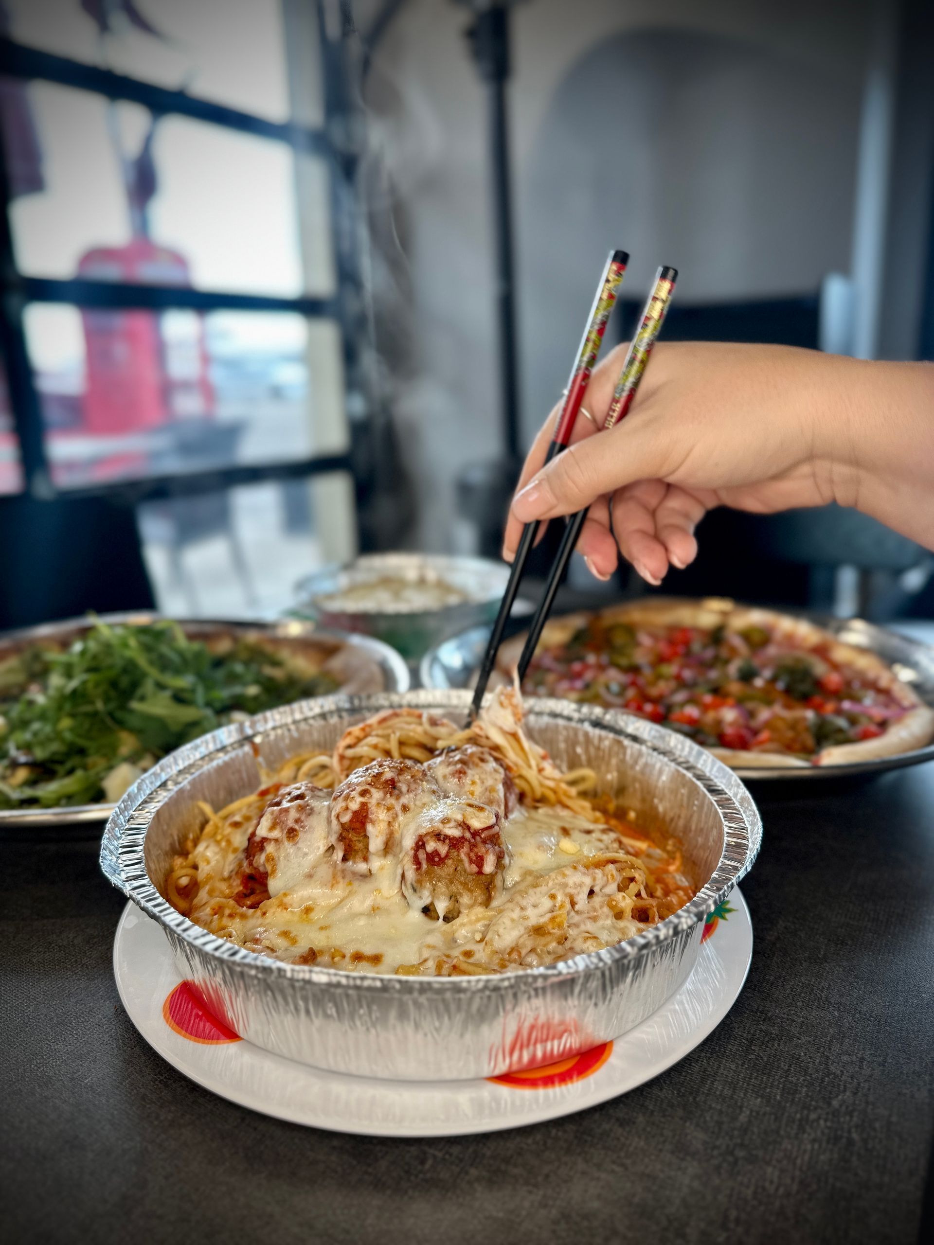 A hand uses chopsticks to lift spaghetti with meatballs and melted cheese from a silver takeout container.
