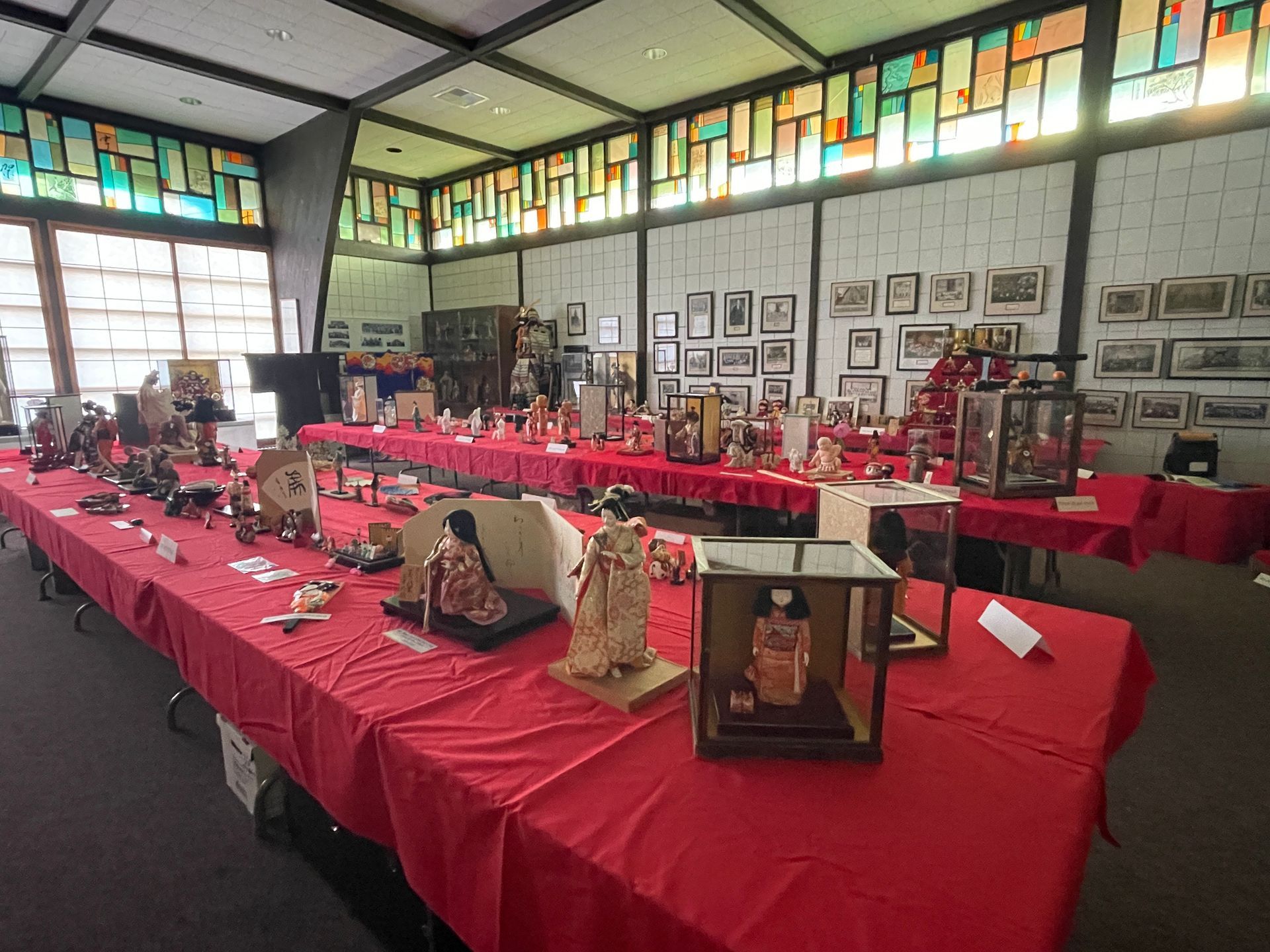 Display of various objects on long tables covered in red cloths, in a room with stained glass windows.