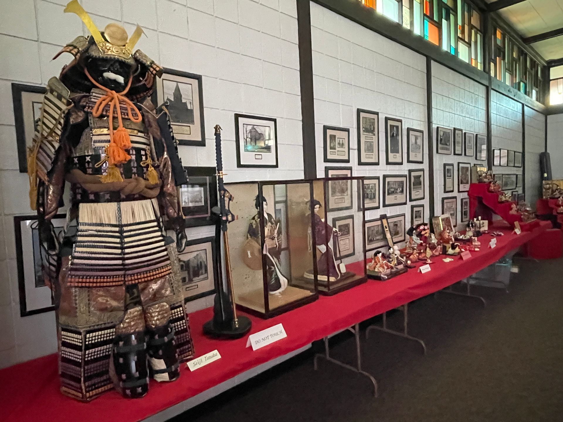 Samurai armor, dolls, and artifacts displayed on a table against a wall with framed pictures and stained glass windows.
