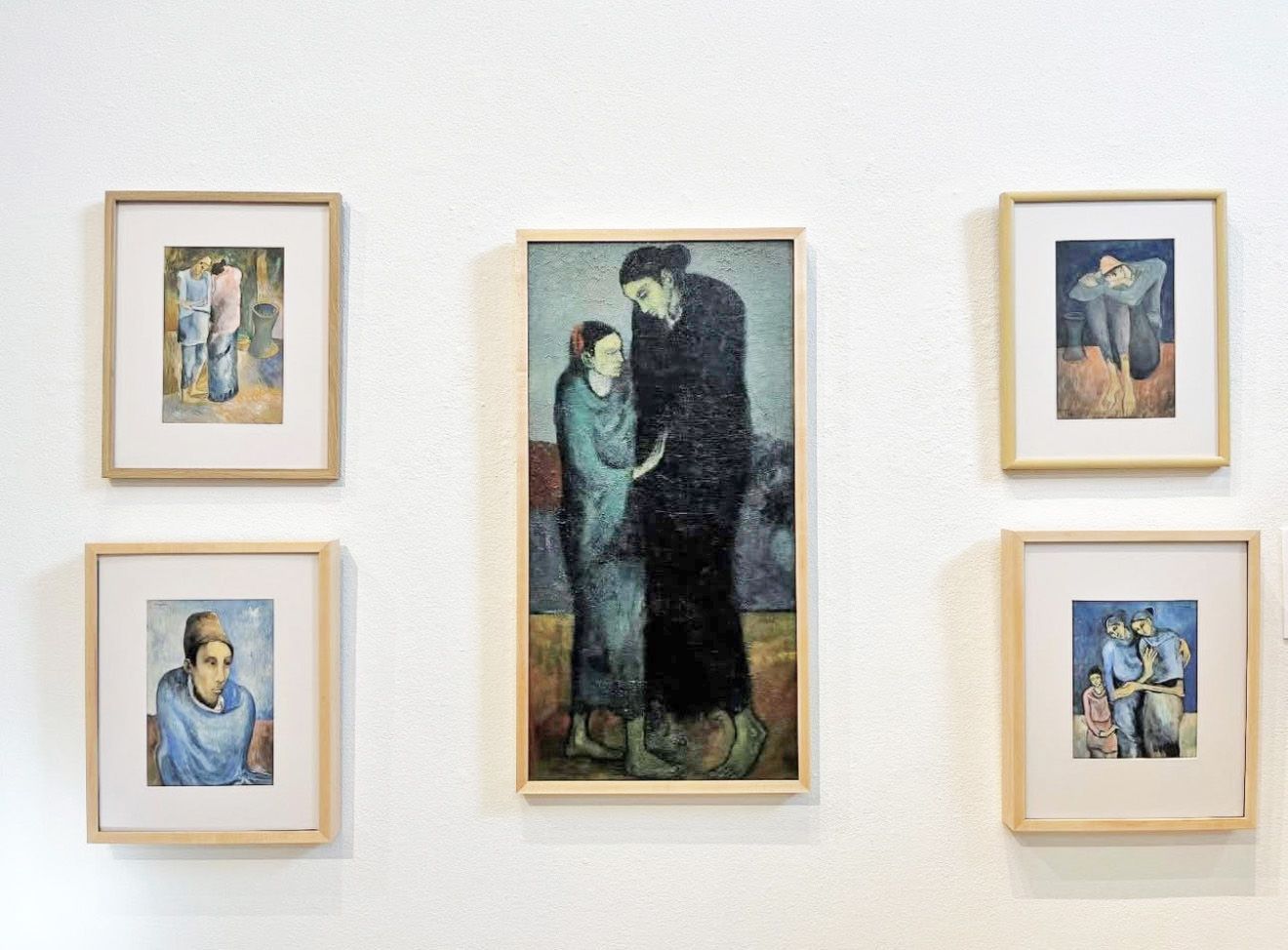 Framed artworks on a white wall; central piece depicts two figures embracing, surrounded by smaller, individual portraits.