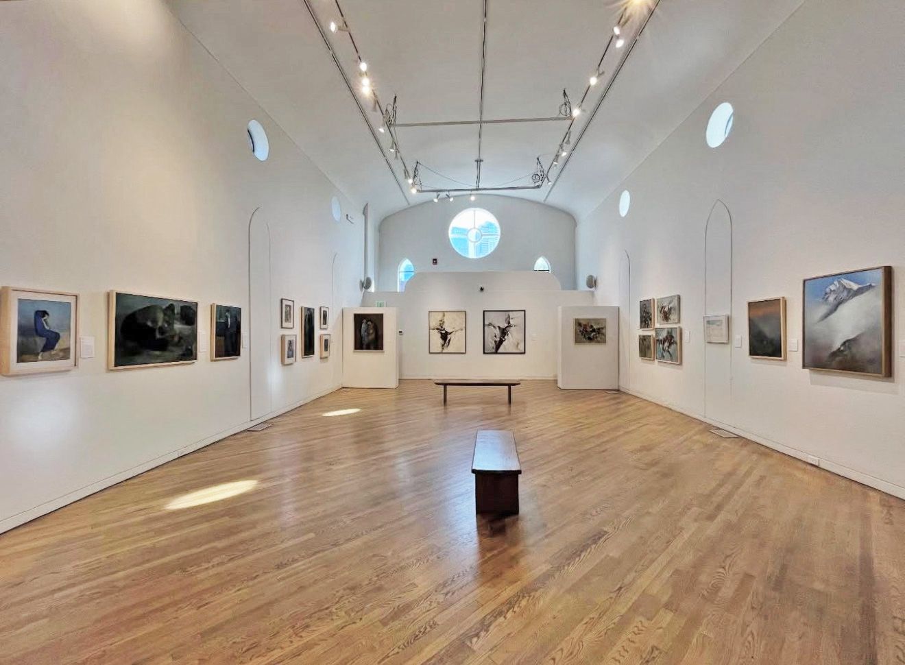 Art gallery interior, white walls, wooden floor, paintings displayed, long room, benches, overhead lighting.