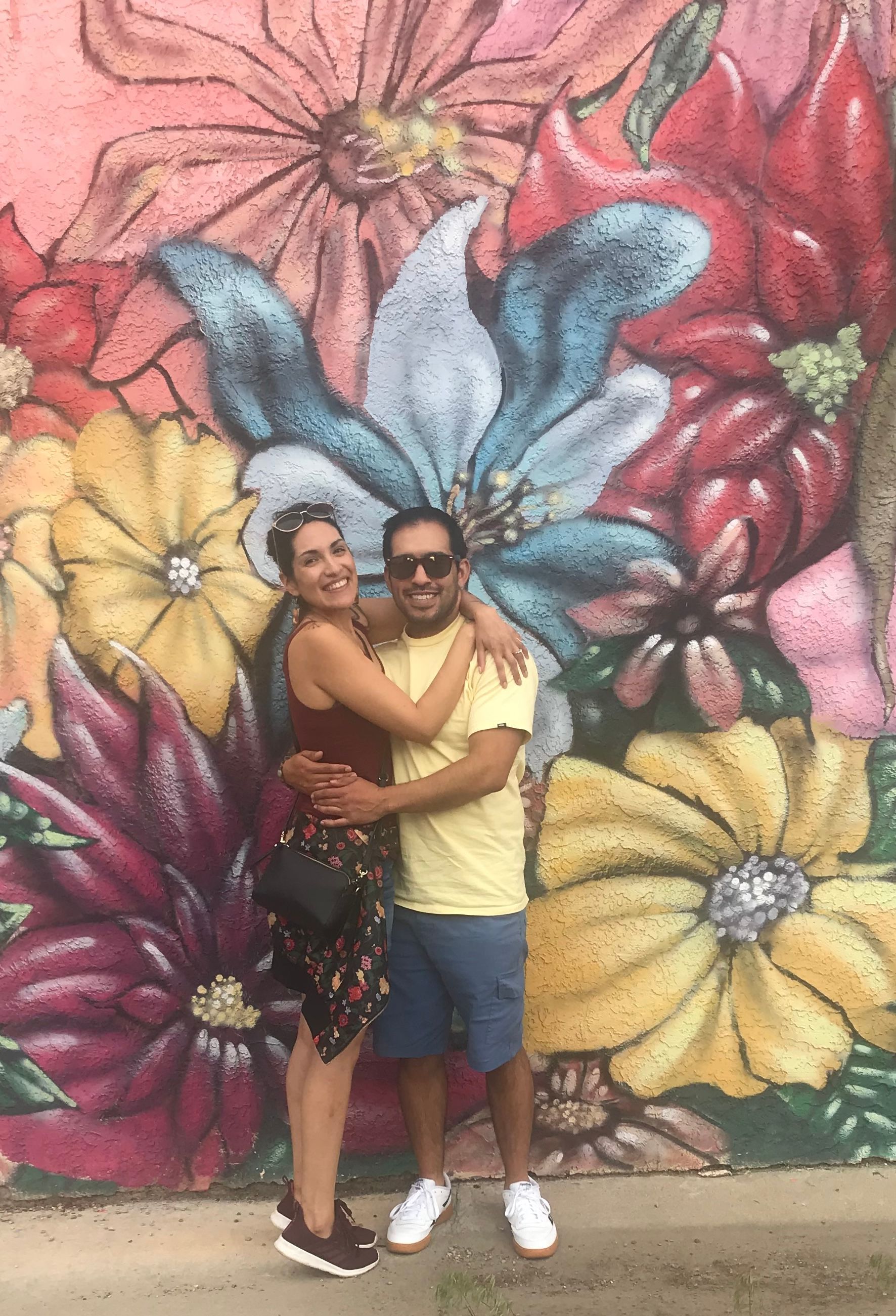 Couple hugging in front of a colorful flower mural. Woman in a floral skirt and top, man in a yellow shirt and shorts.