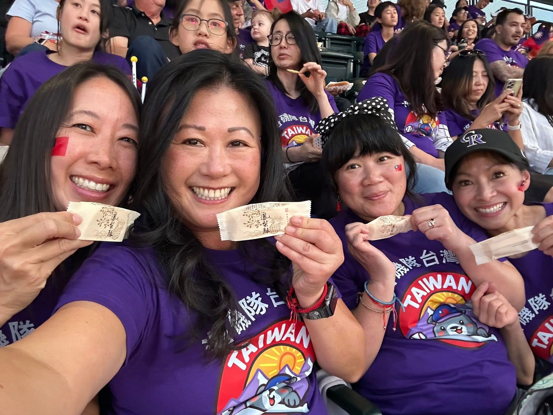 Four people in purple Taiwan shirts hold up paper signs, smiling at a sporting event, with a crowd in the background.