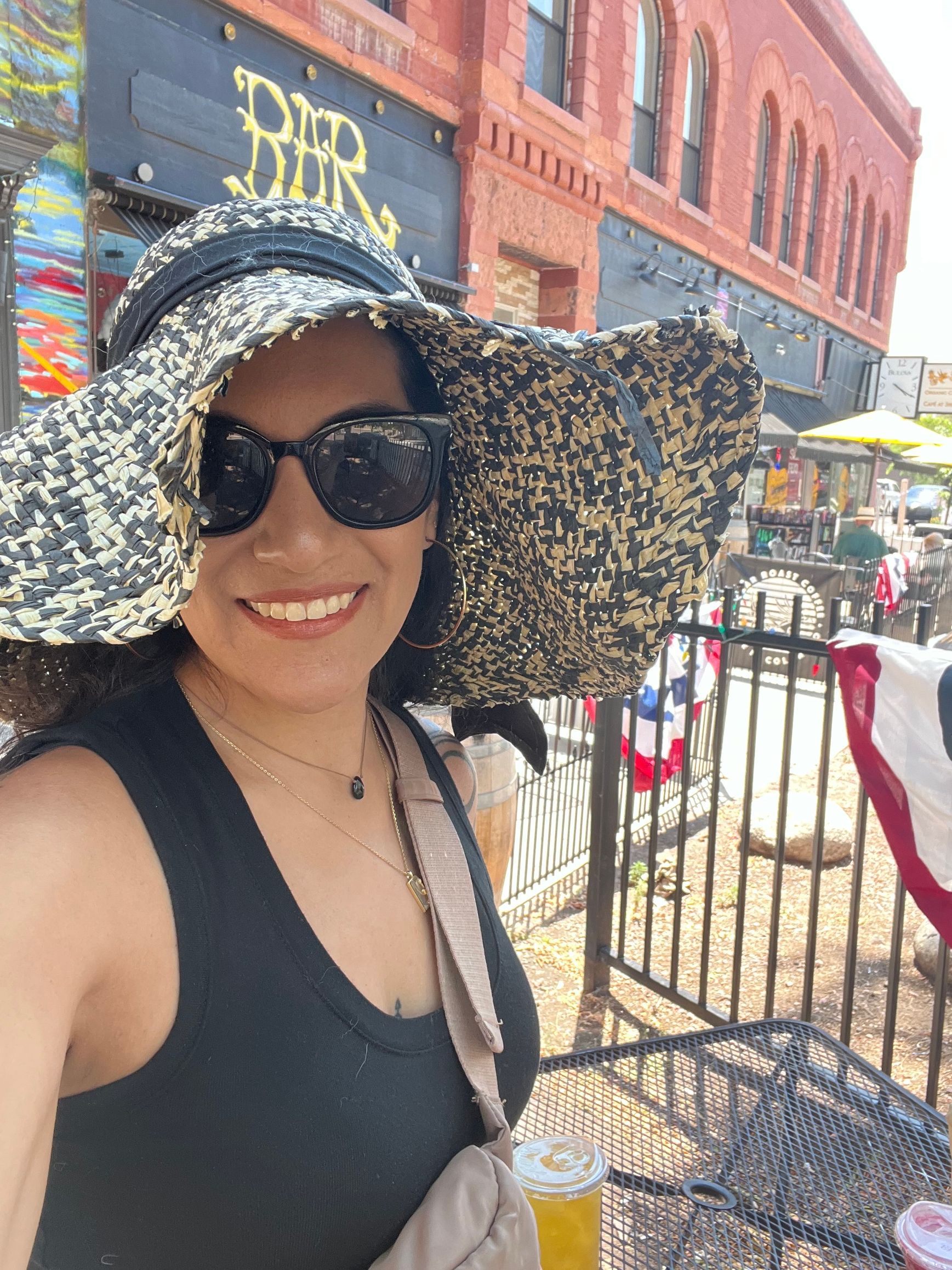 Woman in sunglasses and hat smiles outdoors at a bar, holding a drink.