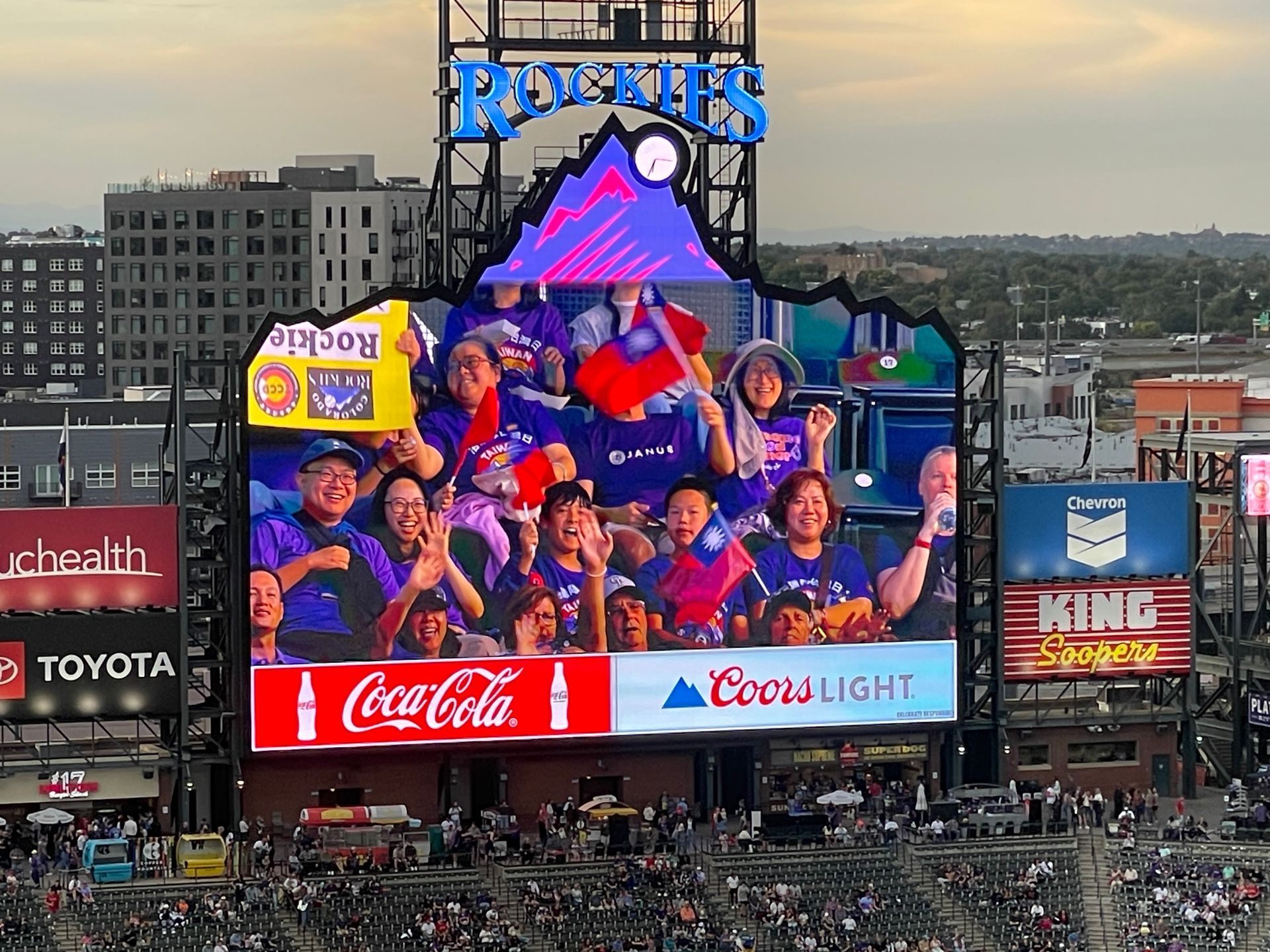 Large scoreboard at Coors Field shows fans cheering. Coca-Cola and Coors Light ads flank it.