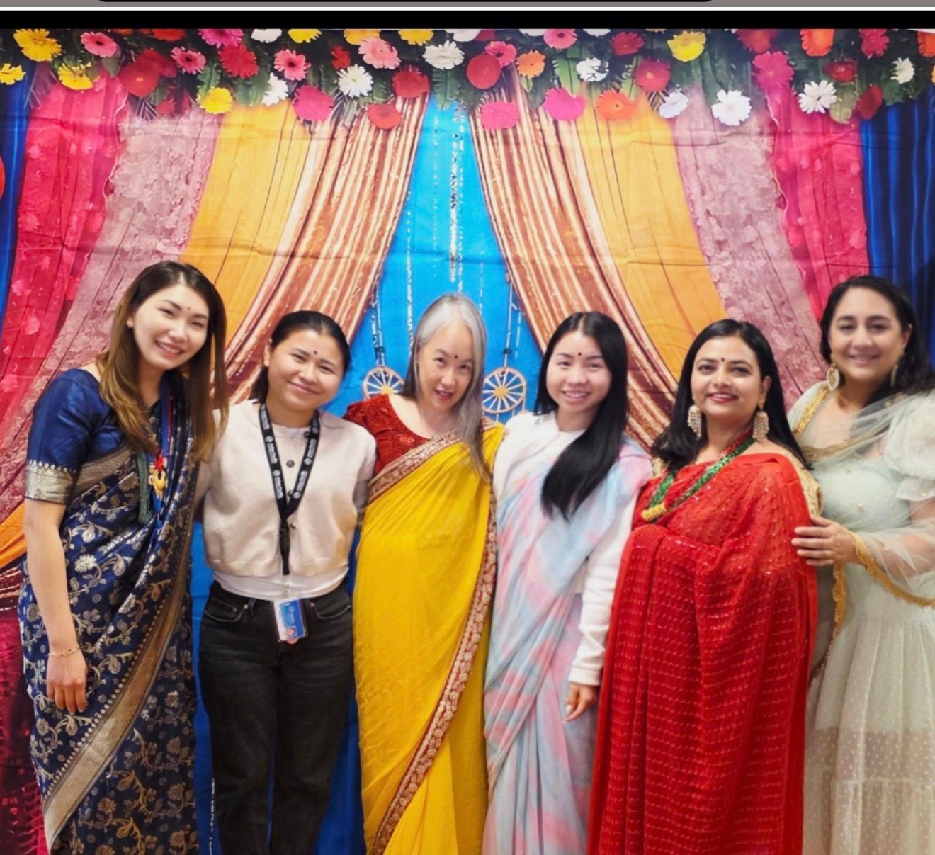 Six women posing in colorful traditional outfits before a bright floral backdrop