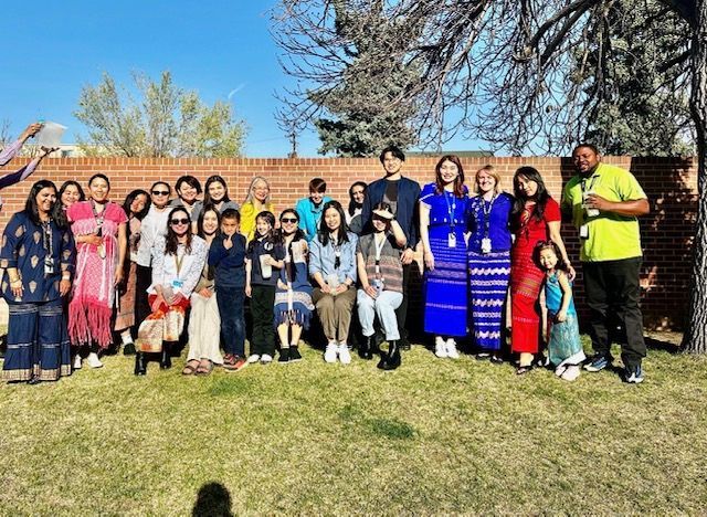 Large group posing outdoors on grass, with trees and a brick wall in the background.