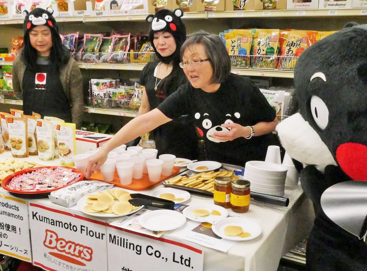 Three people sample snacks at a market booth beside a large black bear mascot.