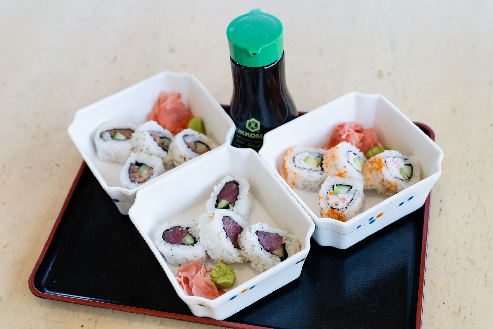 Assorted sushi rolls in white trays on a black tray, with a green-capped bottle behind them.