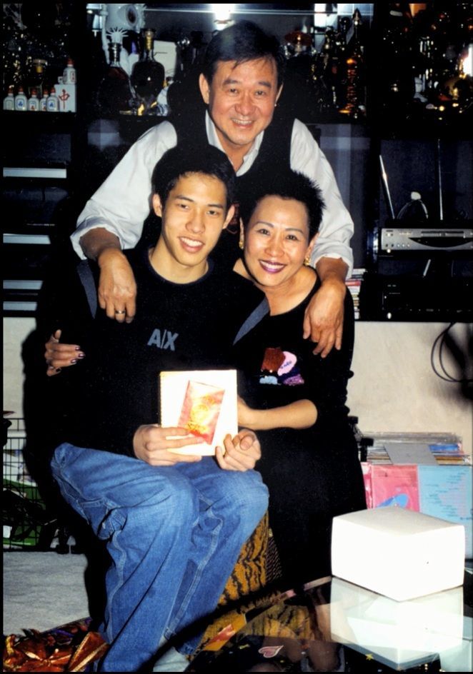 Family of three smiling, posing indoors. Man in vest embraces son, woman with short hair. Son holds small gift.