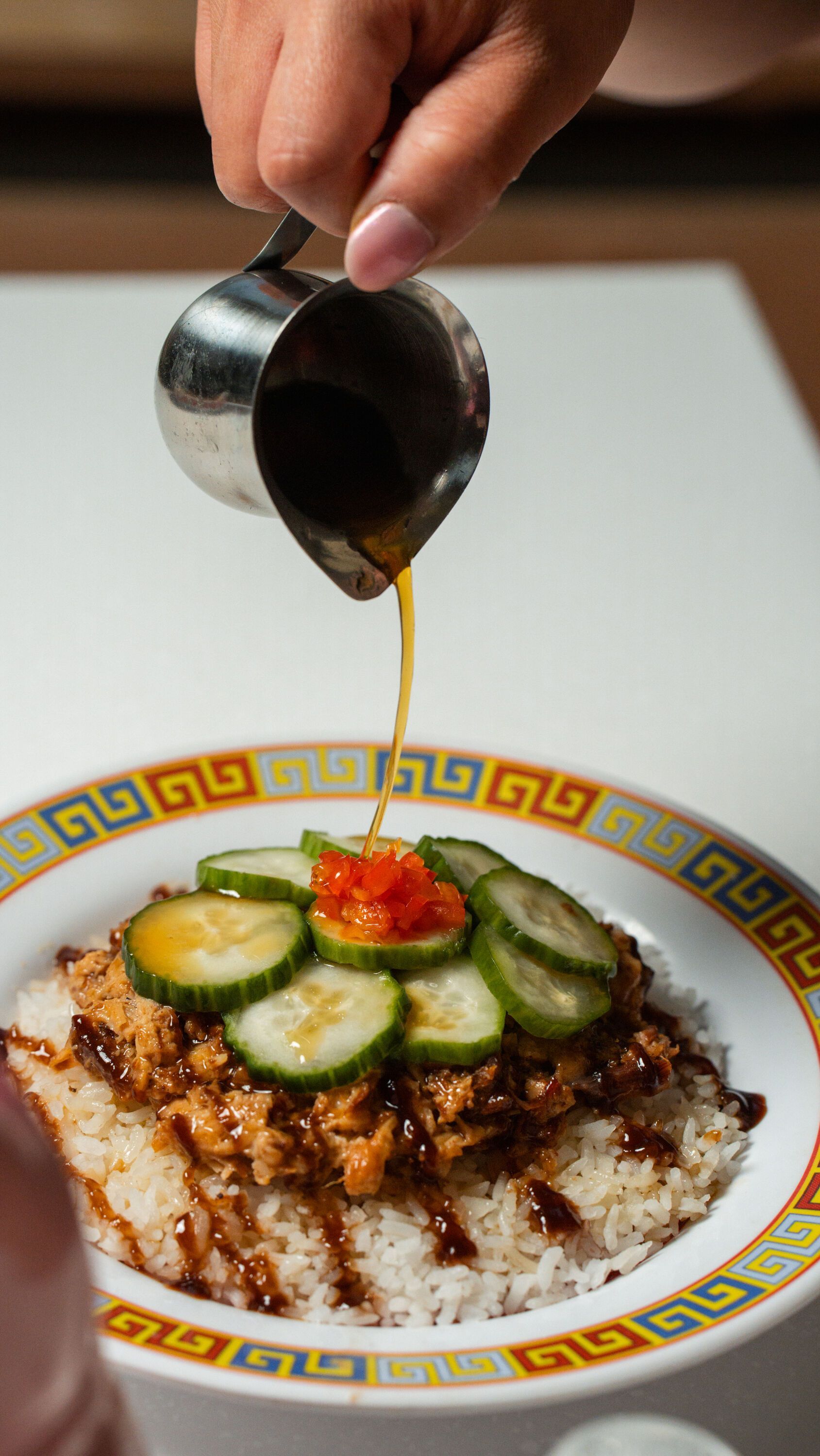 Person pouring sauce over a rice bowl topped with meat, cucumbers, and red relish.