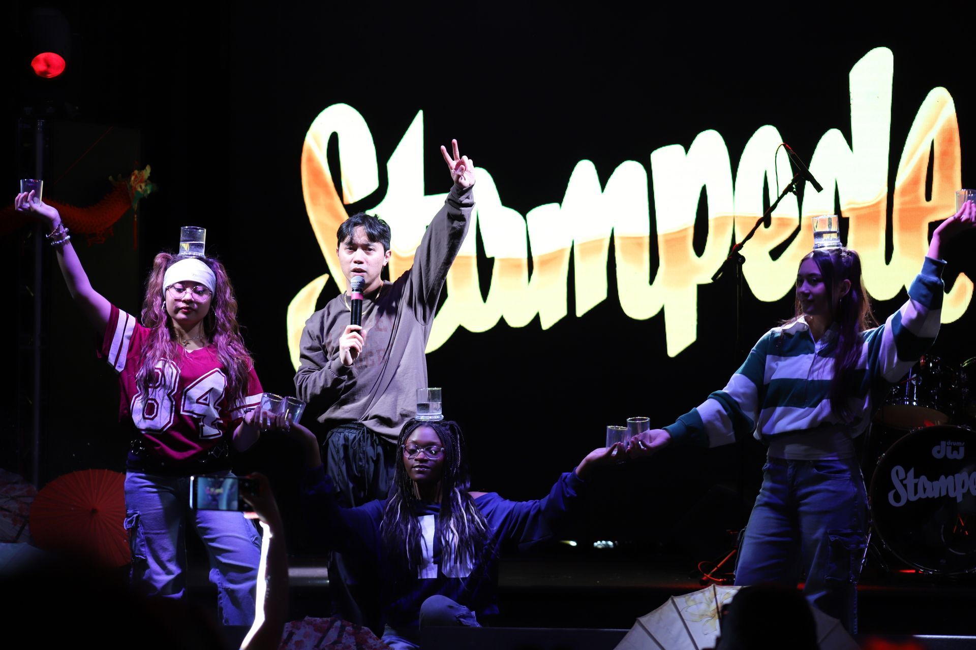 Performers onstage with raised arms in front of a bright “Jumpers” sign.