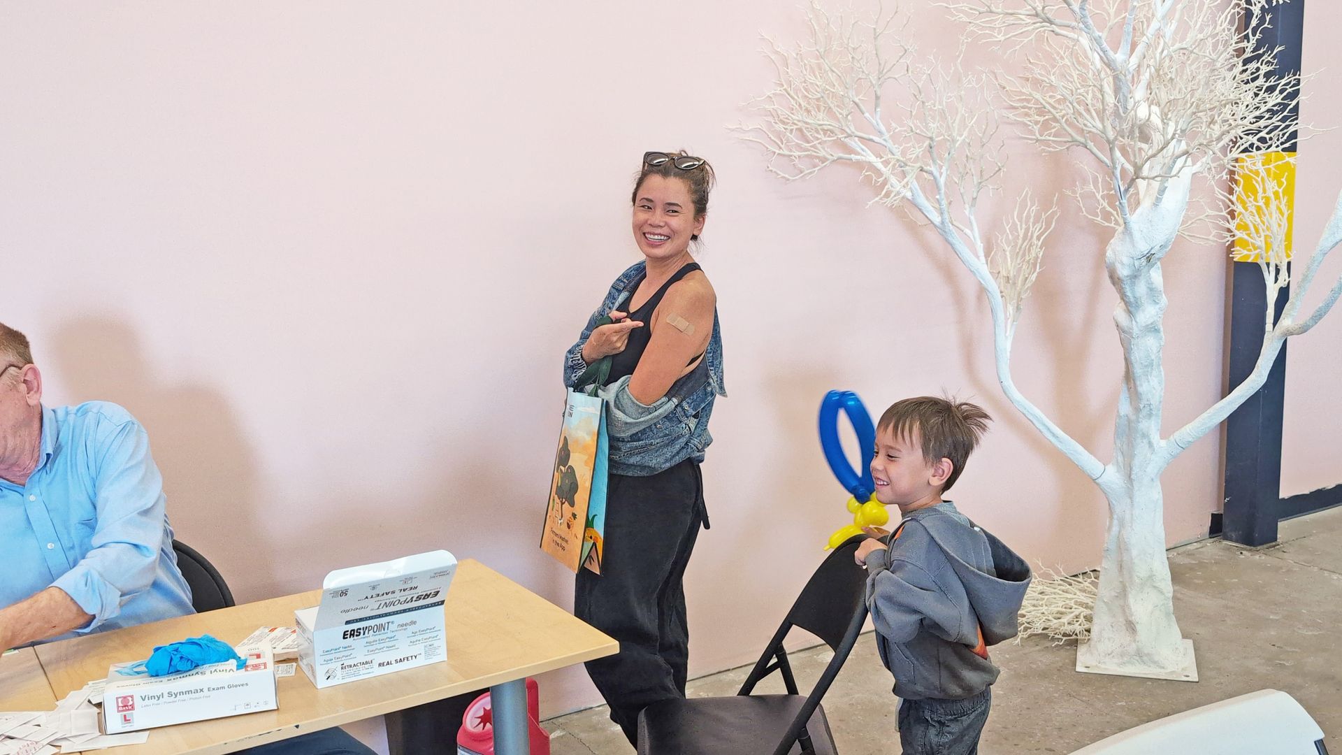 Woman with bandage on arm smiles, child blows bubbles. Pink wall, white tree.