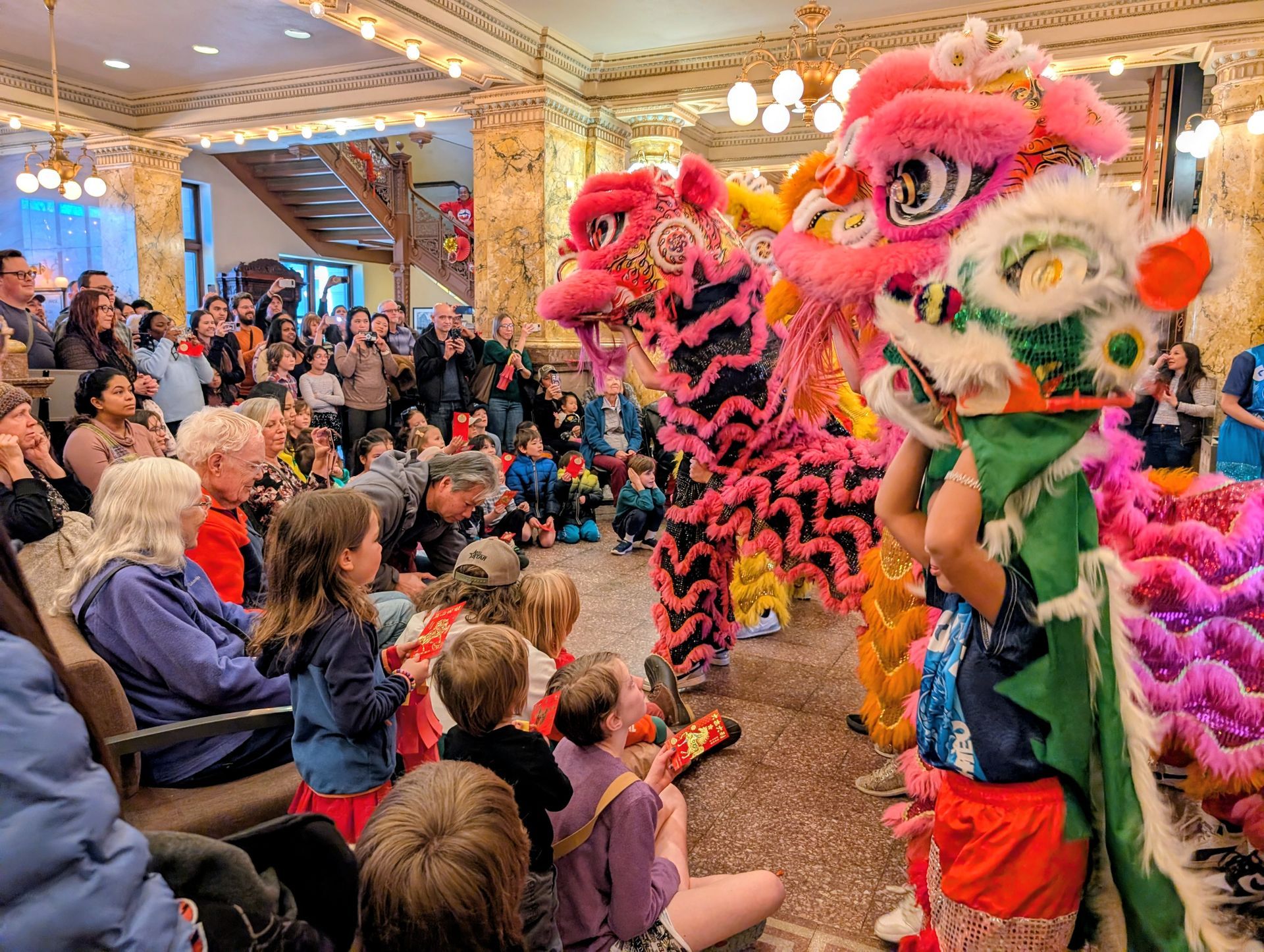 People gather in an ornate building to watch performers in colorful, elaborate lion dance costumes.