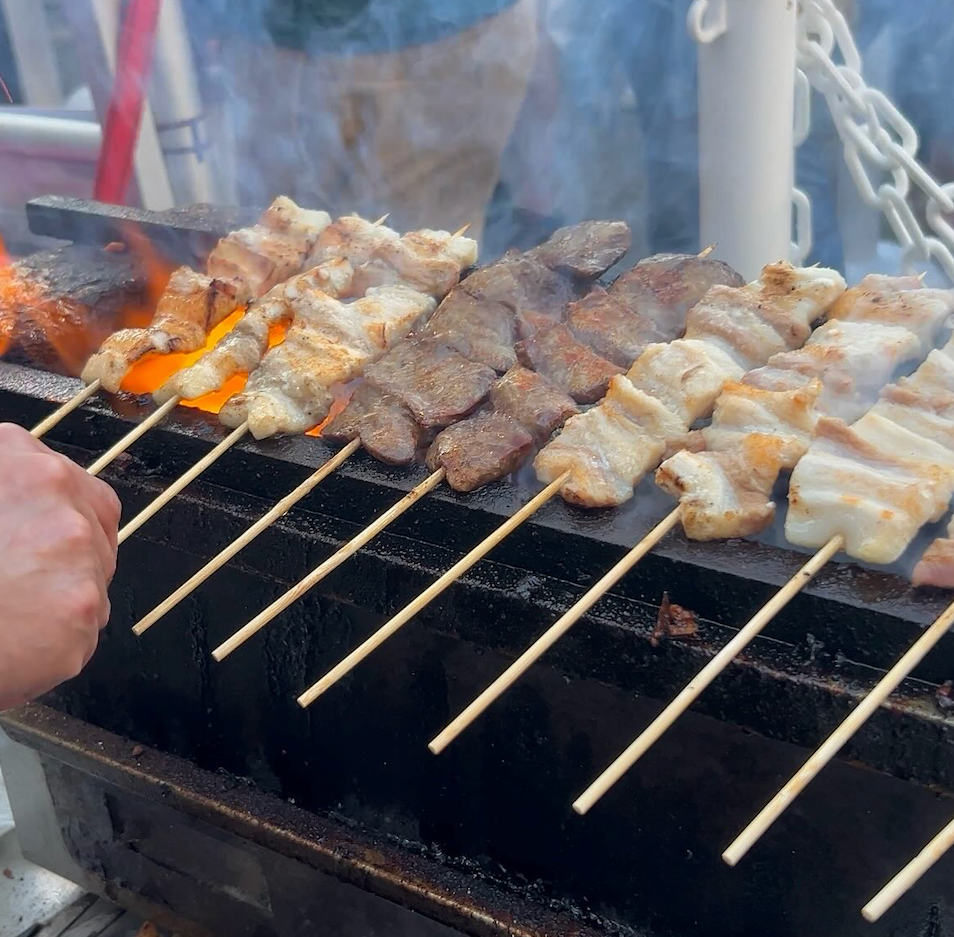 Skewered meats grilling over flames. A hand turns the skewers. Outdoor setting, smoke rising.