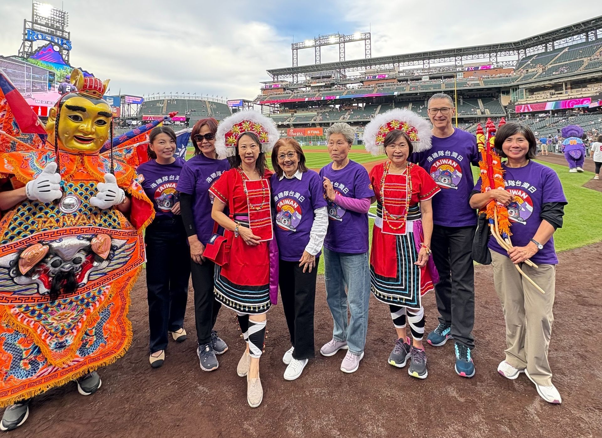Group of people in traditional attire and purple shirts pose on a baseball field with a costumed figure.