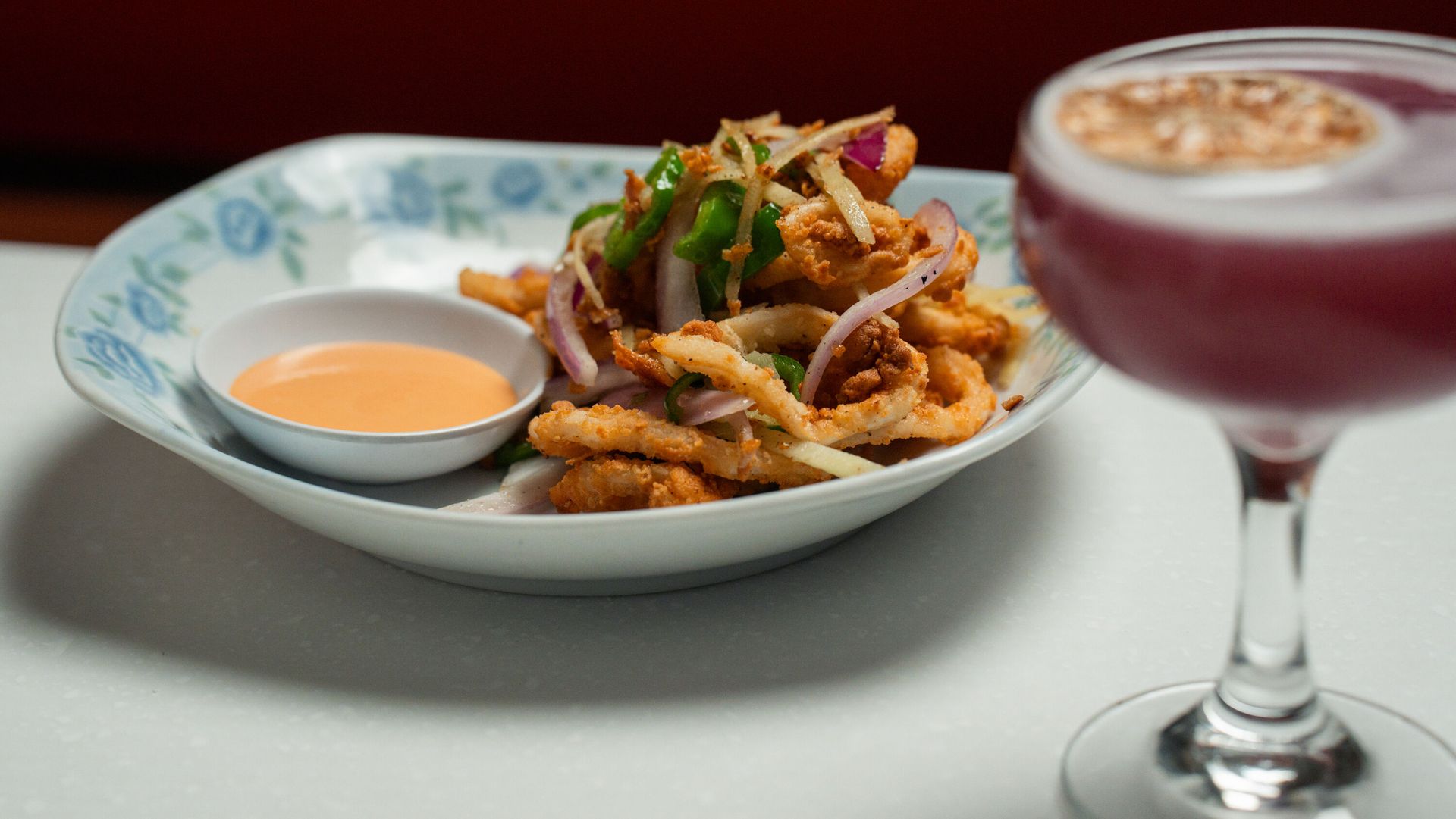 Fried calamari in a floral bowl with dipping sauce, and a purple cocktail, on a white table.