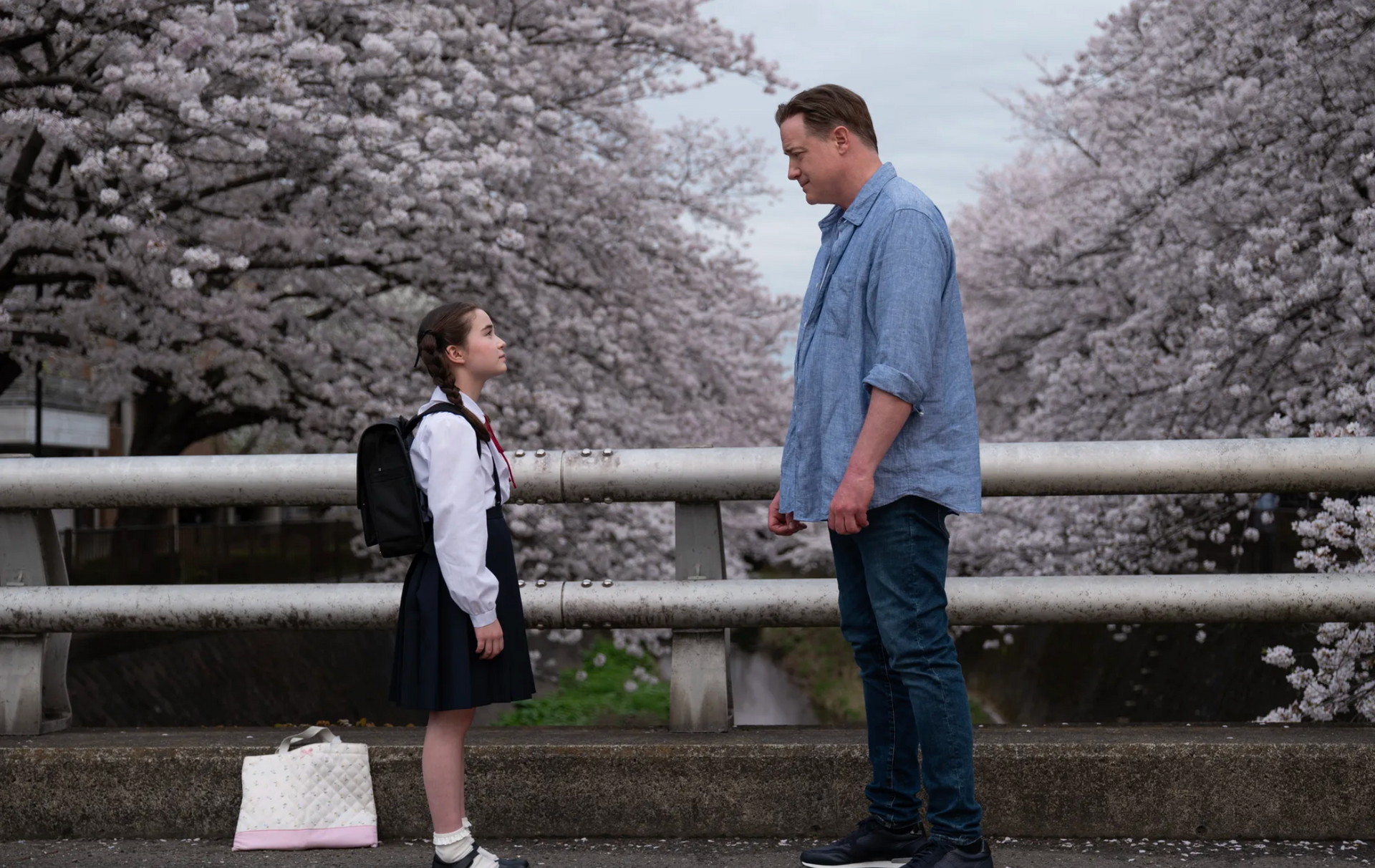 A person stands face-to-face with a child on a bridge lined with cherry blossom trees.