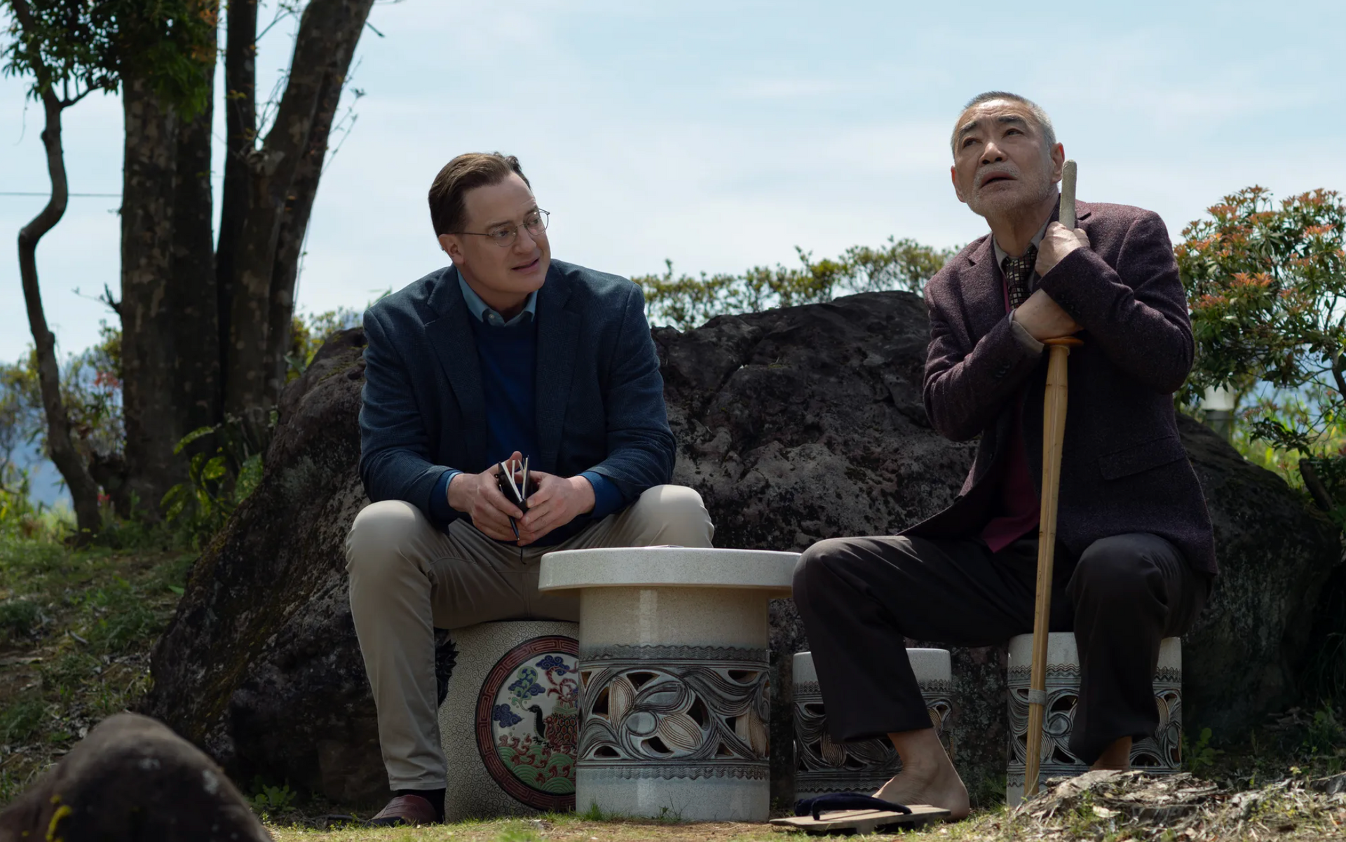 Two men seated outdoors, conversing. One holds a staff. Sunny day, greenery in background.