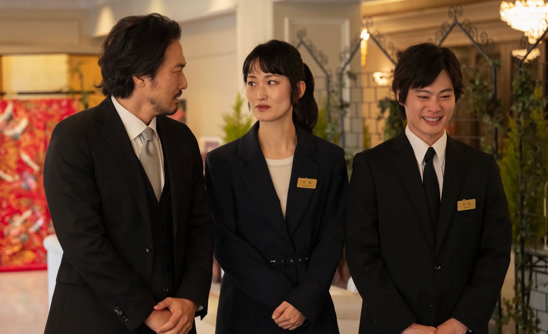 Three people in formal attire stand indoors; two look at the third, who is speaking. Setting appears to be a reception hall.