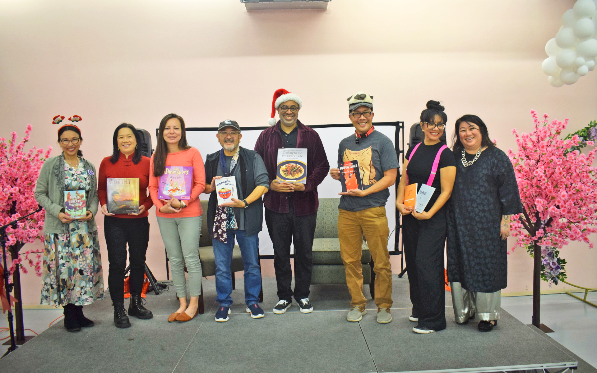 Group of people holding books on stage. Pink and white decor, some wearing hats.