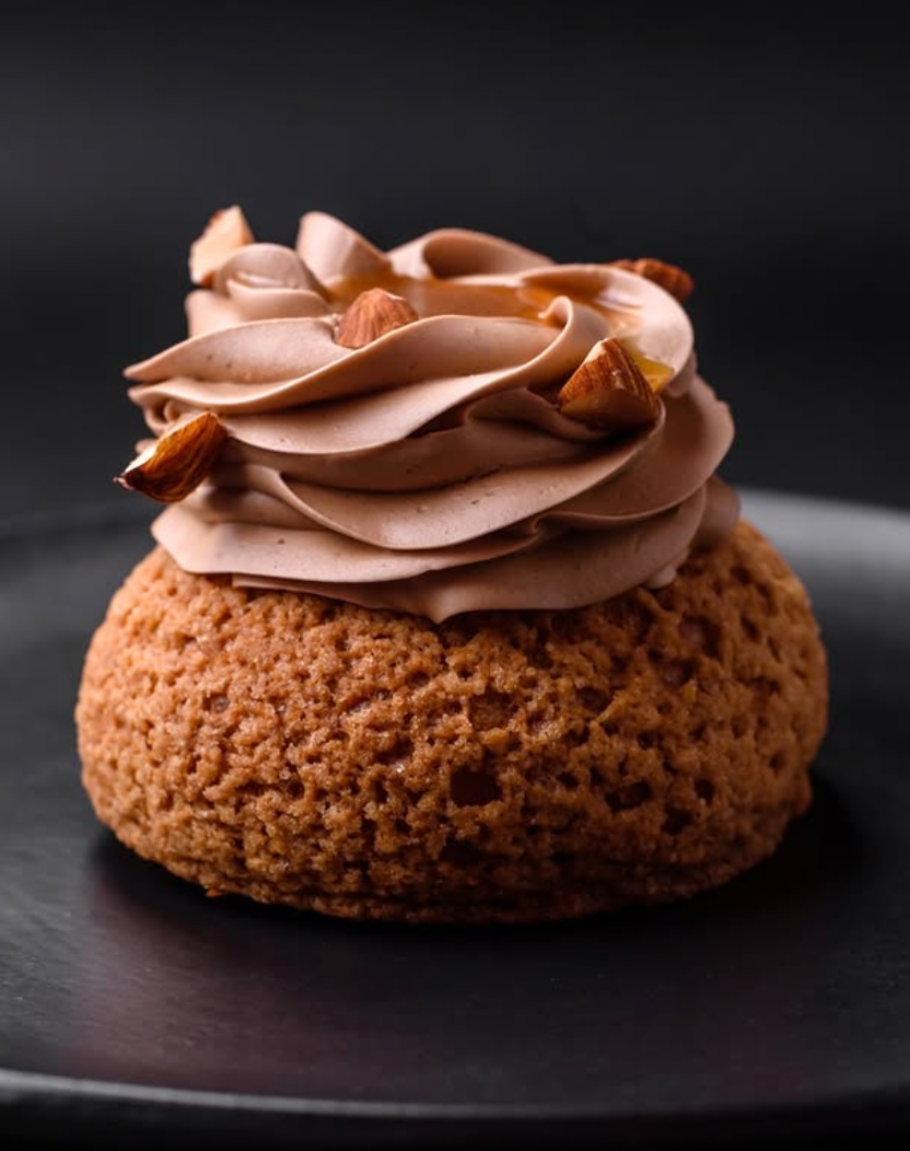 A single choux pastry topped with a swirl of light brown cream and toasted nut pieces, centered on a matte black plate.