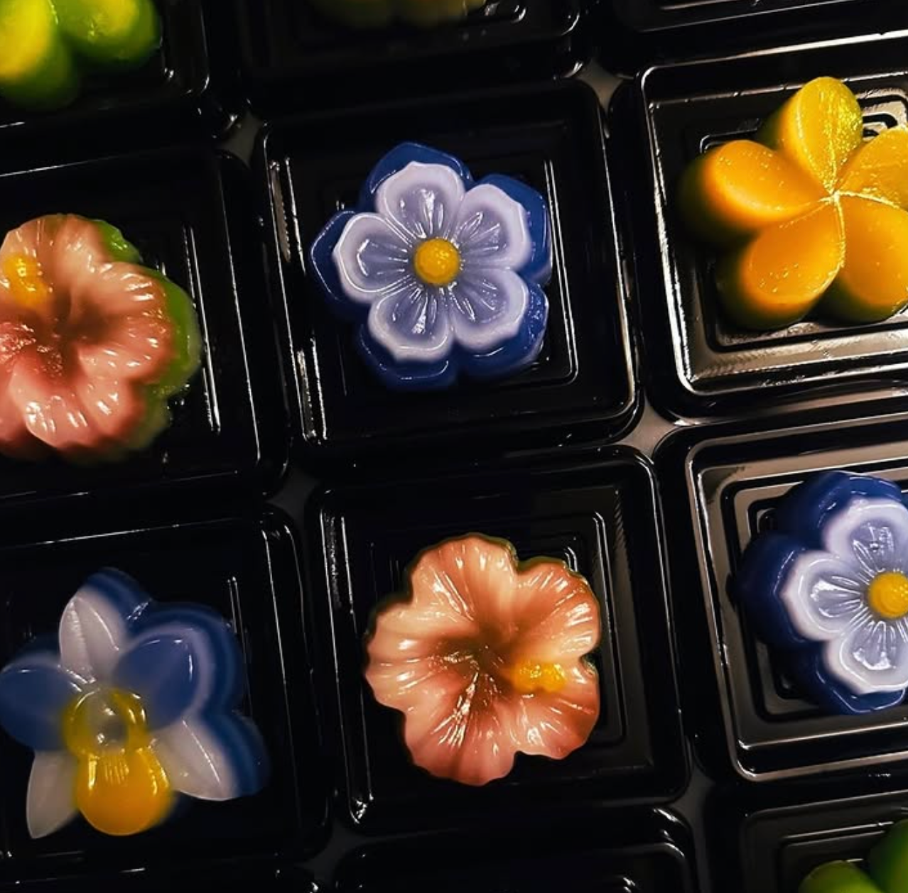 Colorful, flower-shaped Thai jellies served in individual black plastic compartments.