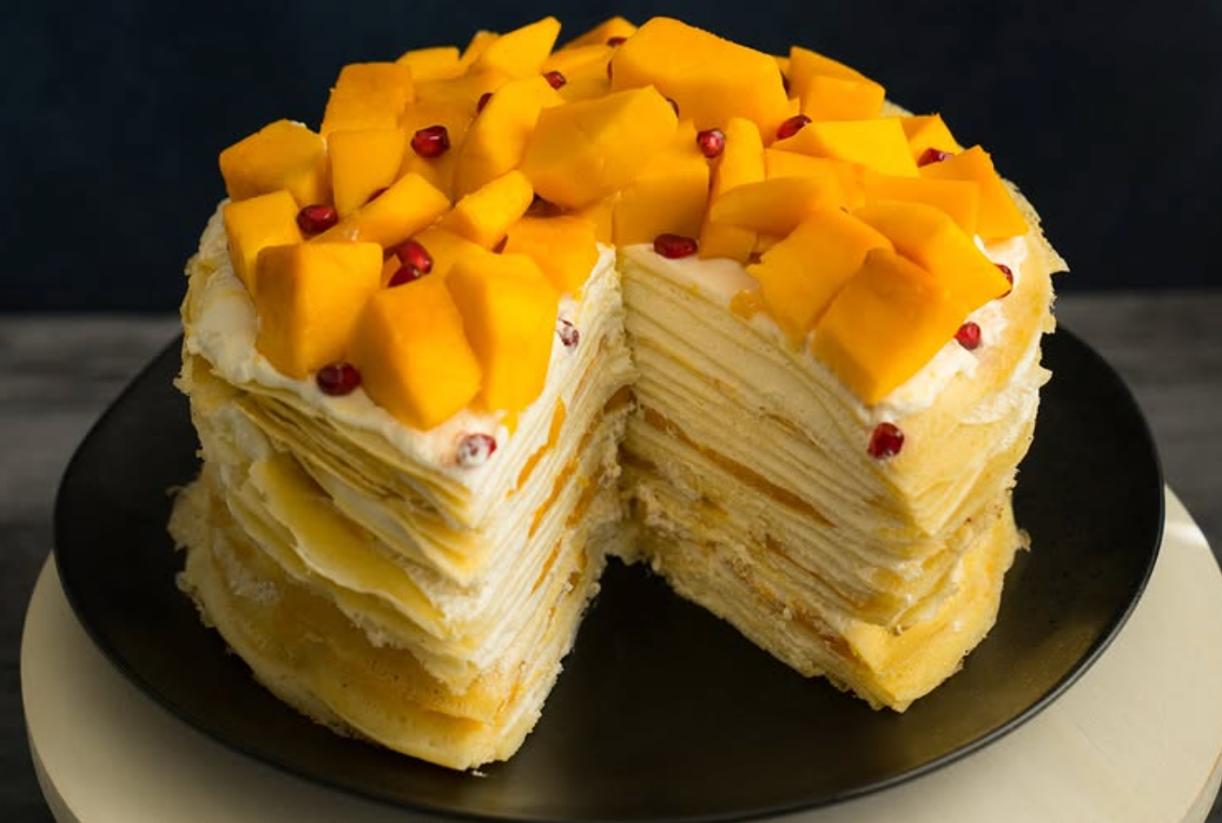A mango mille-crepe cake topped with fresh mango cubes and pomegranate seeds sits on a black plate against a dark background.