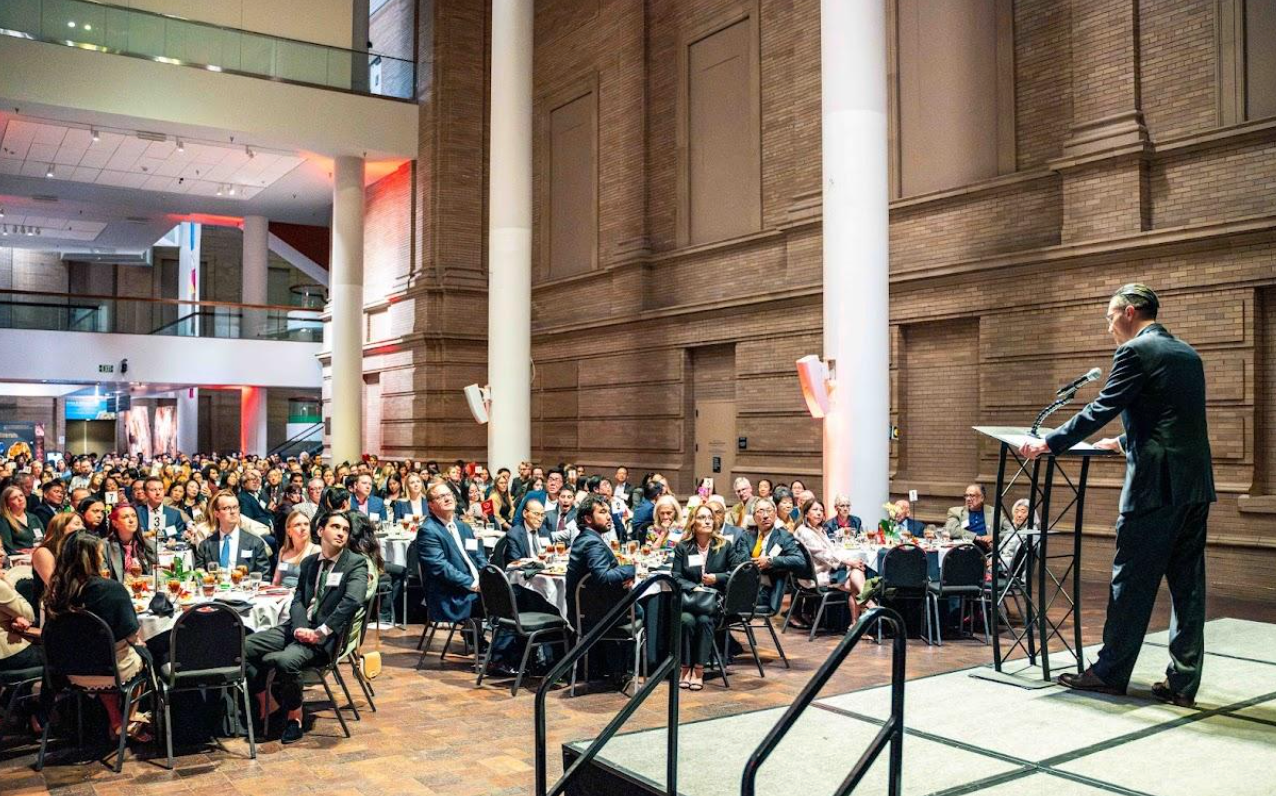 Speaker addressing seated audience in a large auditorium with brick walls and stage lighting