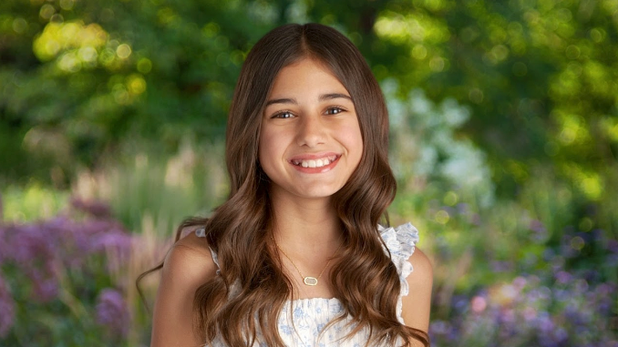 Girl with long brown hair smiles, wearing a white dress, against a blurred garden background.