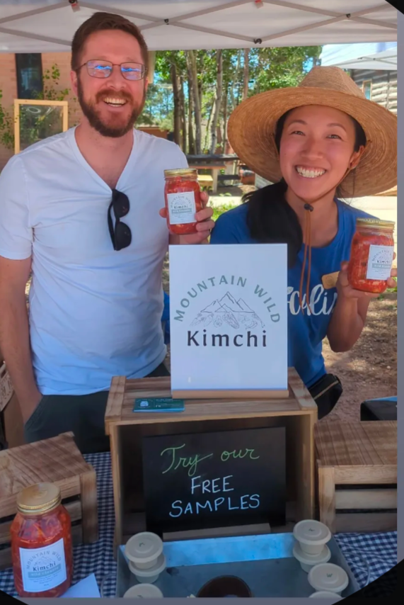 Two people smiling at a farmer's market, holding kimchi jars next to their product sign.