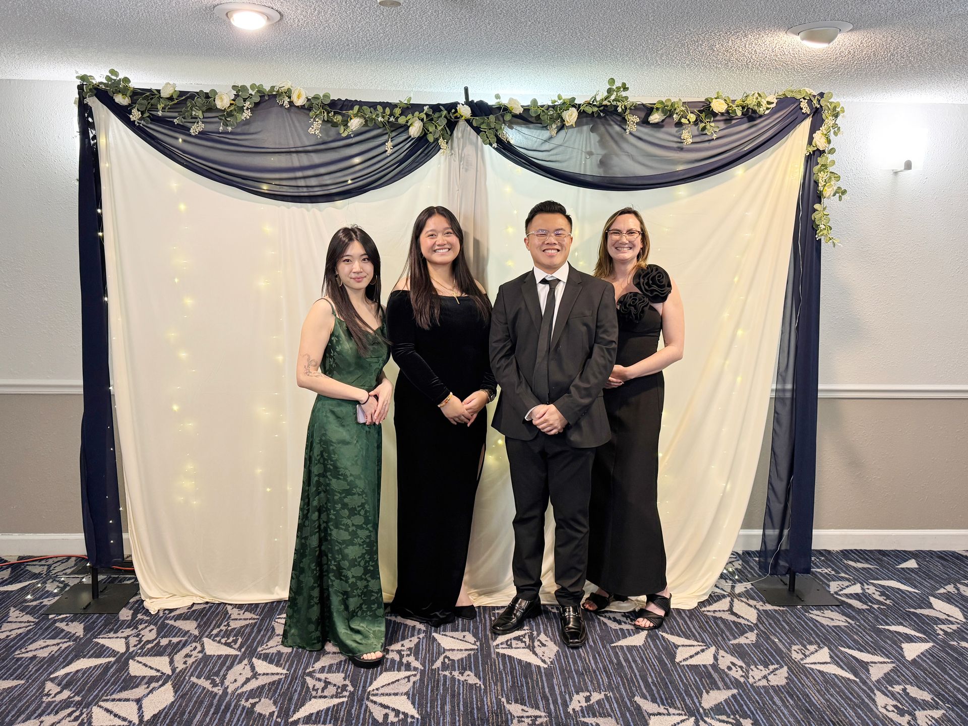Four people pose formally in front of a draped backdrop; one in green dress, three in black attire.