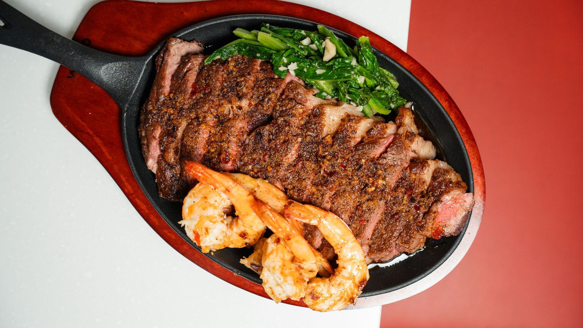 Sliced steak, shrimp, and greens served on a sizzling platter, red background.