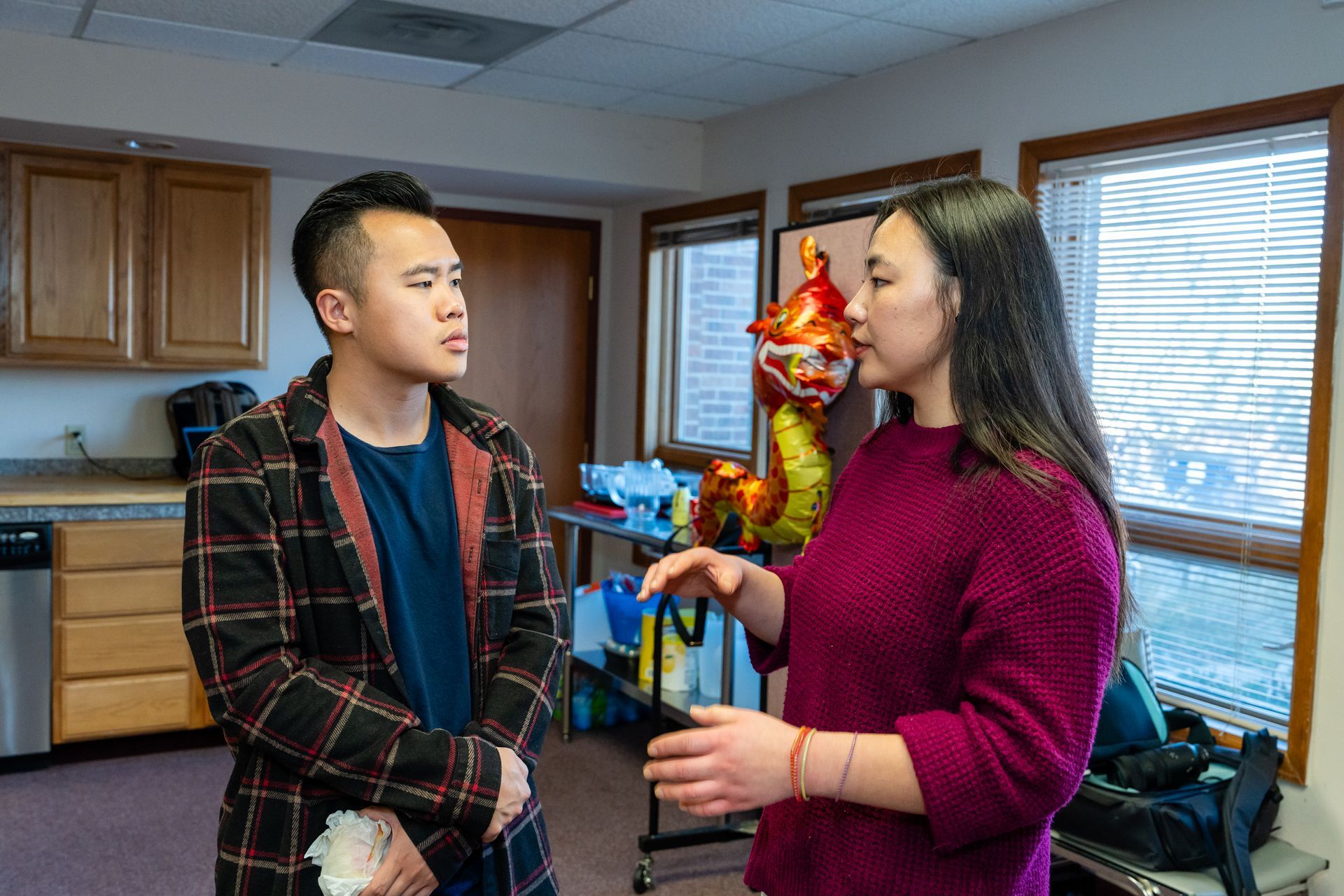 Man in plaid coat listens as woman in purple sweater gestures, in room with cabinets and balloons.