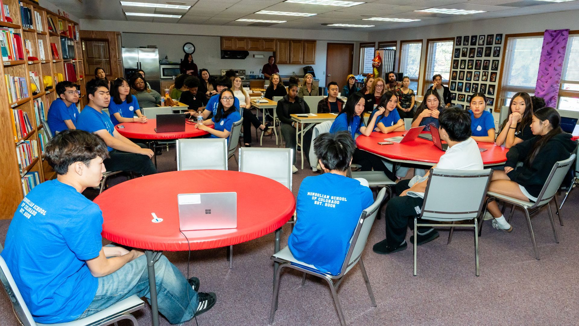 People in blue shirts and others at round tables with laptops, in a room with windows and bookshelves.