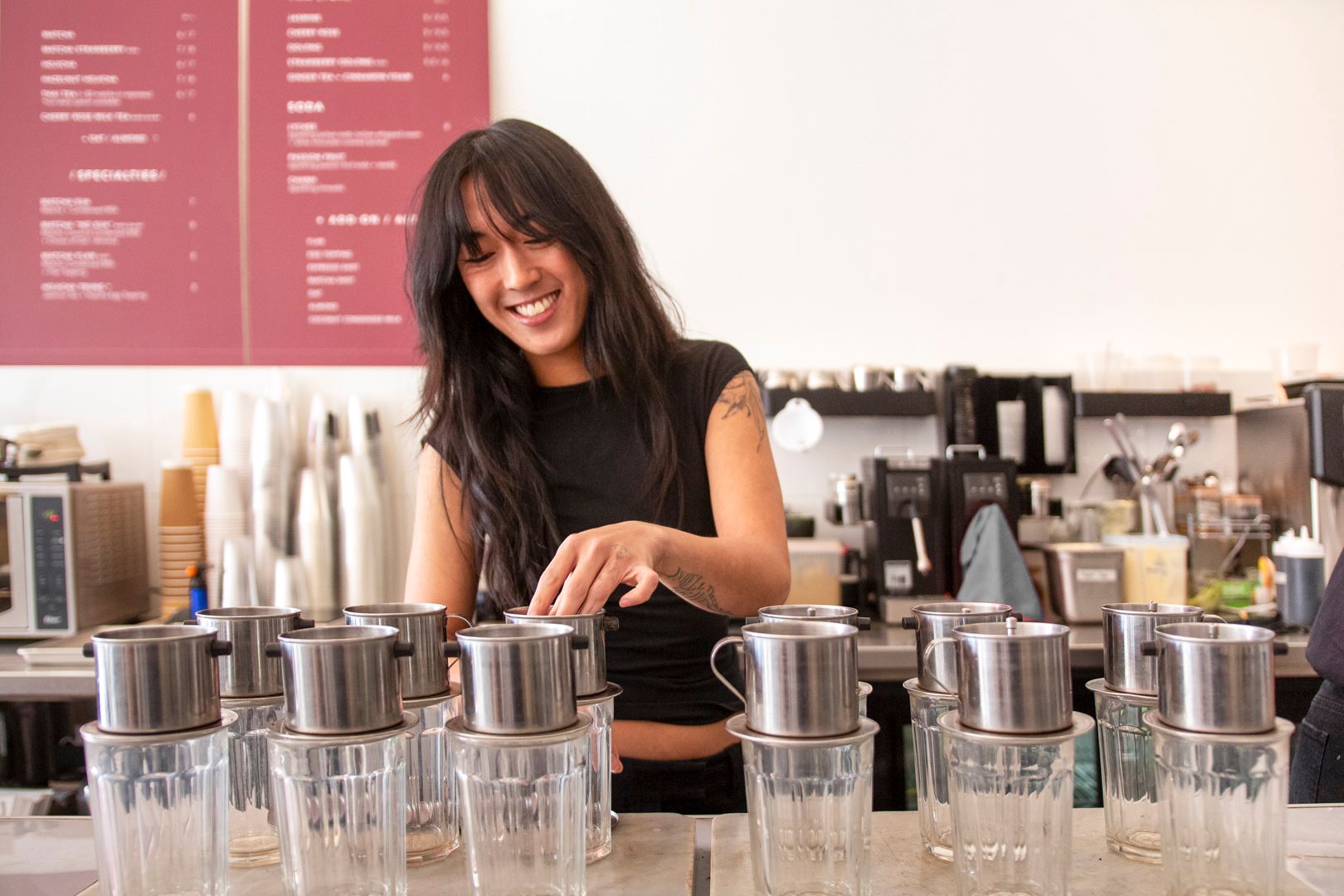 A smiling person in a black shirt prepares coffee using metal Vietnamese drip filters on glass cups in a cafe.