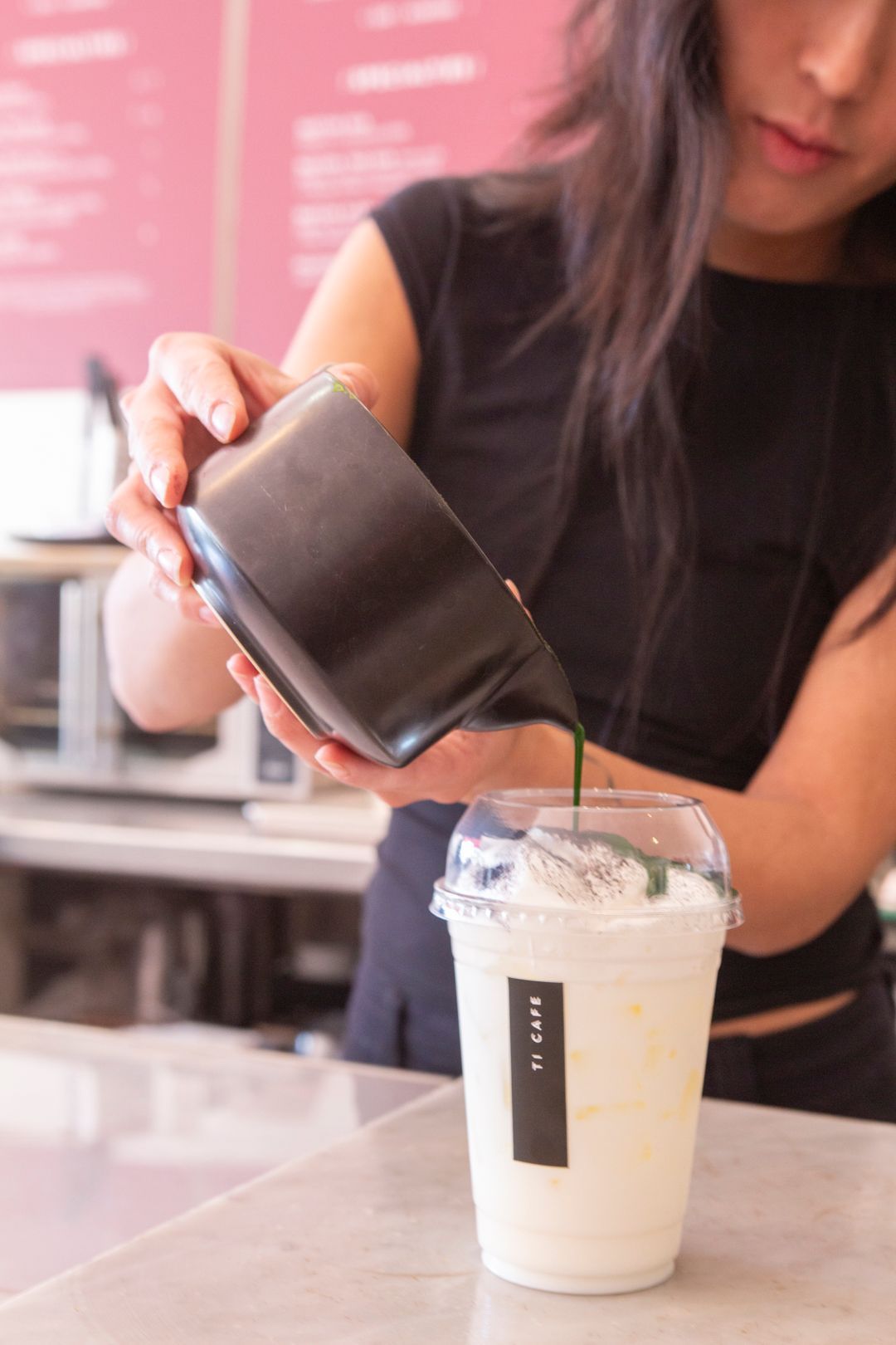 A person pouring green matcha tea from a dark pitcher into a plastic cup filled with milk and ice at a cafe counter.