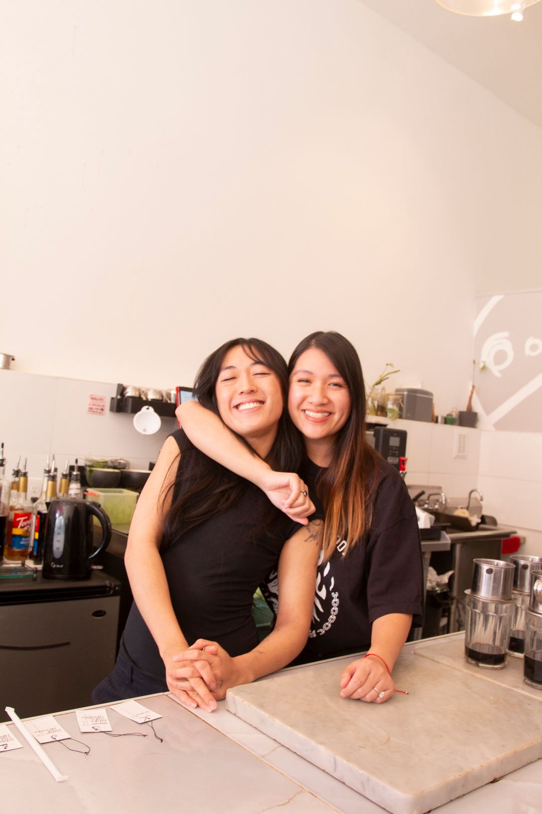 Two smiling people with long dark hair embrace behind a café counter with coffee equipment and a stone surface.
