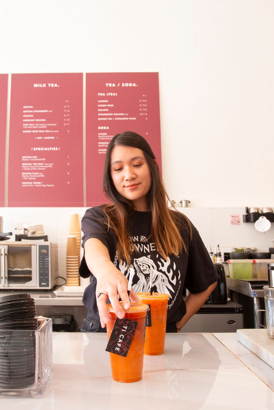 A person sets an orange boba tea drink on a counter in a shop with a menu board visible on the wall behind them.