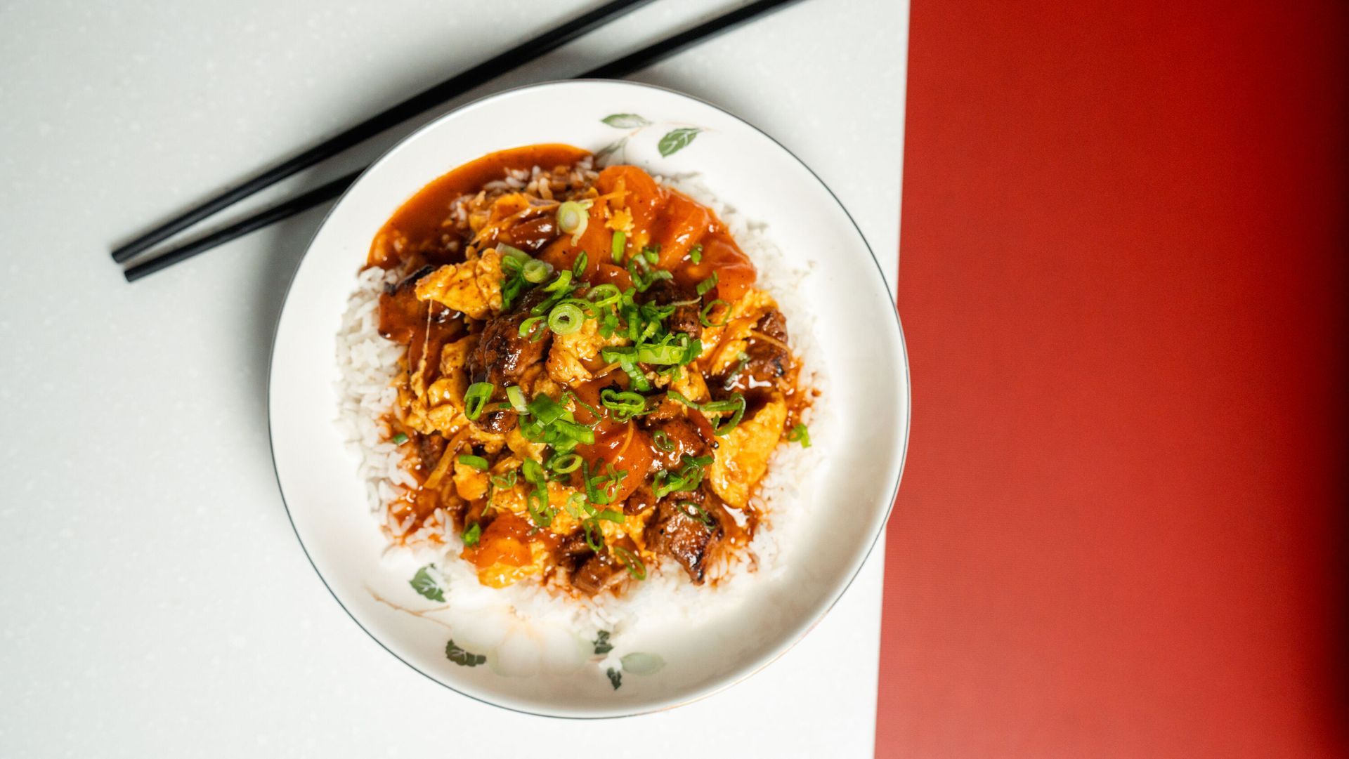 Plate of food: rice with sauce, meat, and green onions, served with chopsticks on a white and red background.