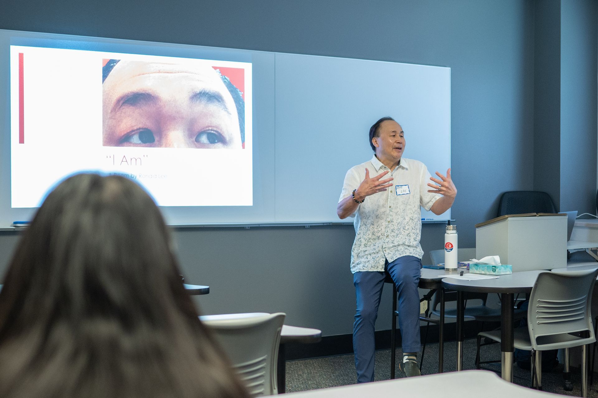 A man giving a presentation in a classroom, gesturing with hands. Eyes on screen, student in foreground.