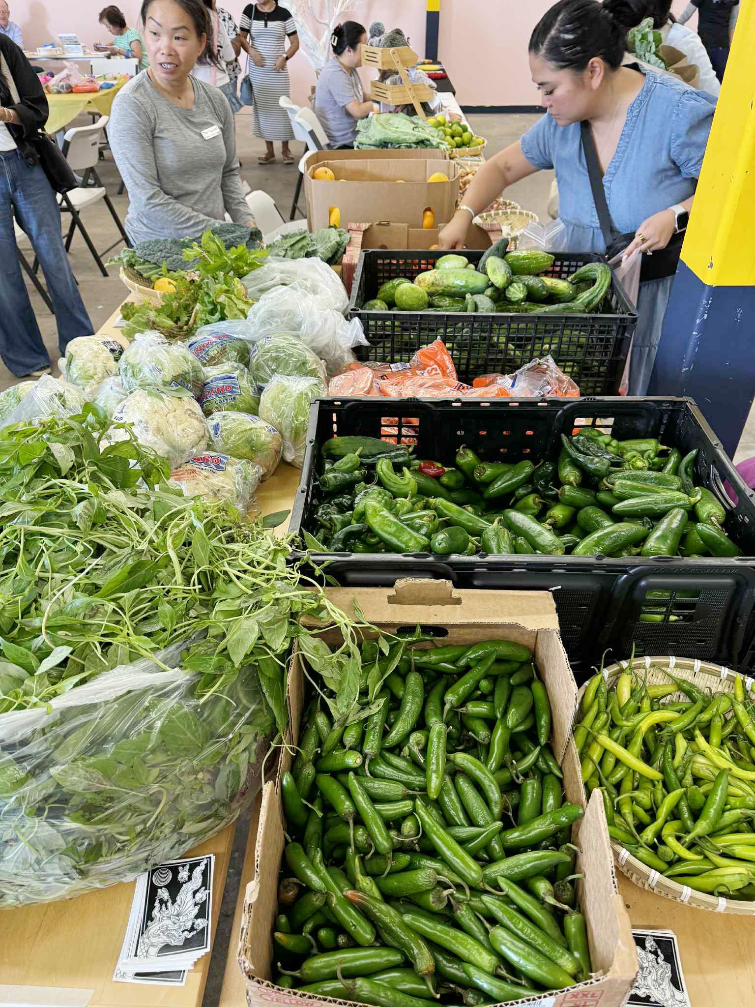 Produce stand at an indoor market. Shoppers browse bins of green peppers, cucumbers, and cauliflower.