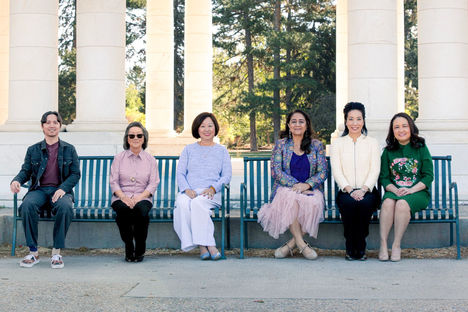 Six people seated on a bench in front of white columns and trees, outdoors in daylight.
