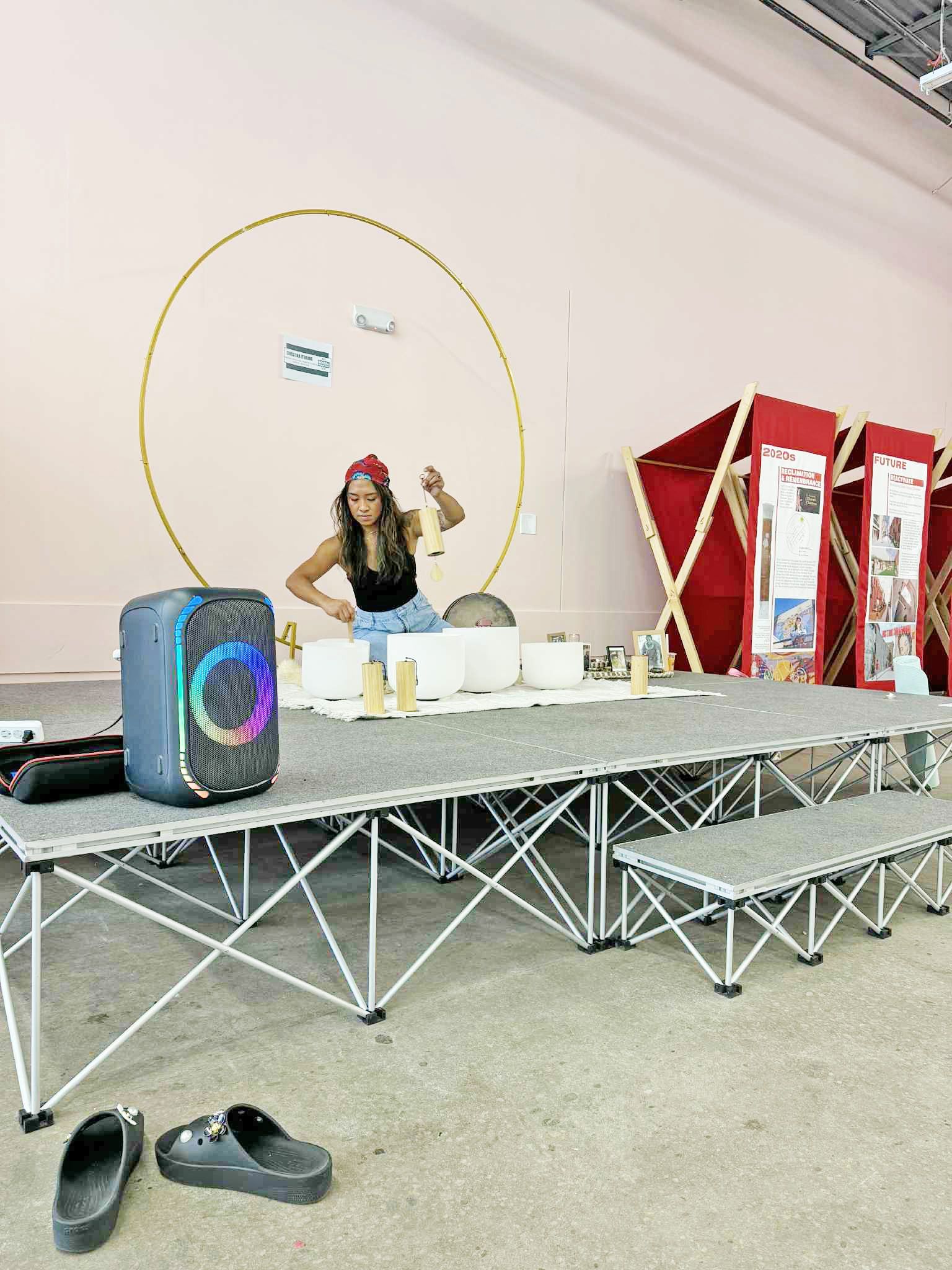 Woman on stage smiles near a speaker, backdrop of a gold hoop and structures.