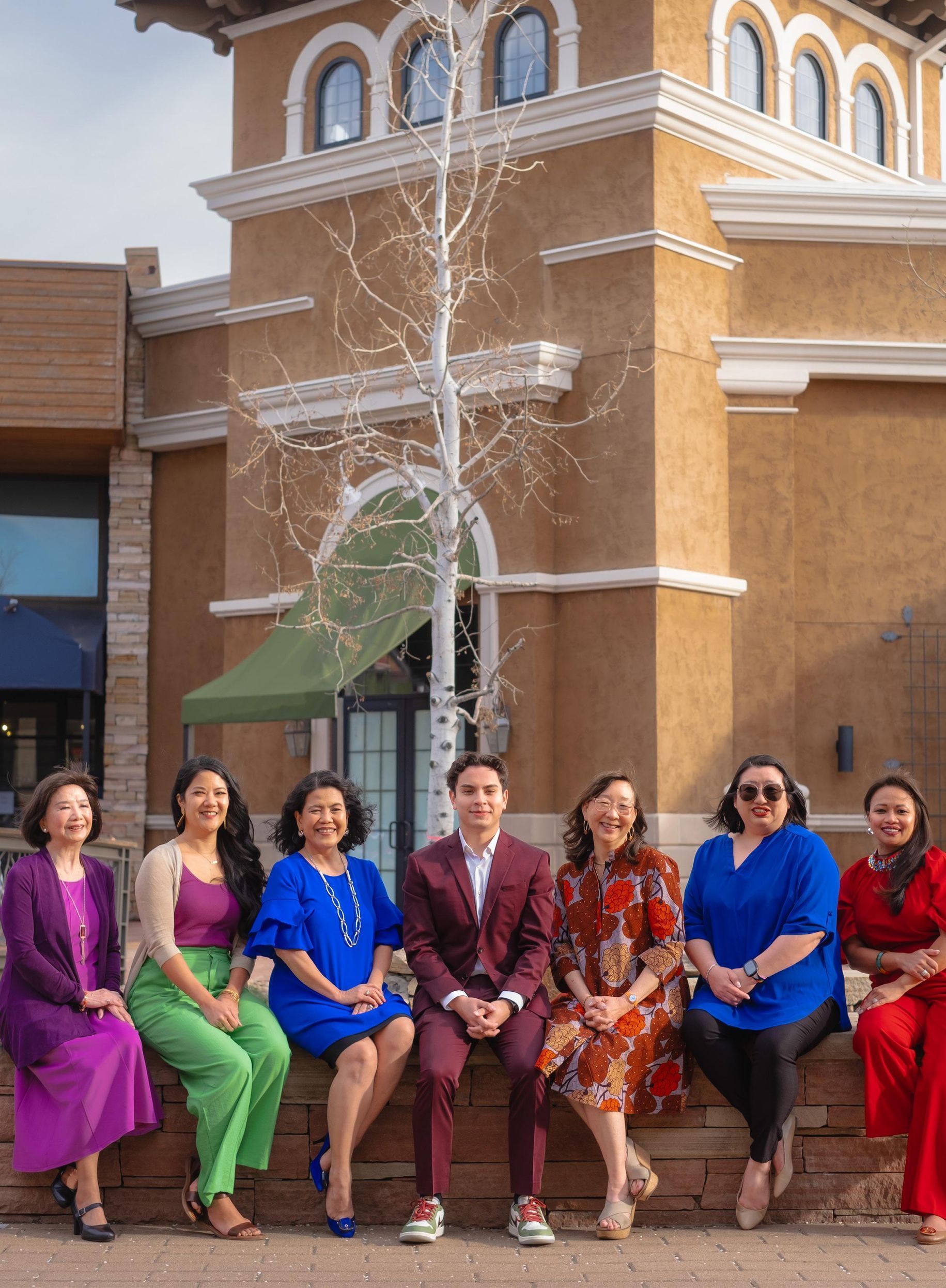 Group of people sitting outside a tan building. Colorful clothing: purples, blues, greens, and reds.