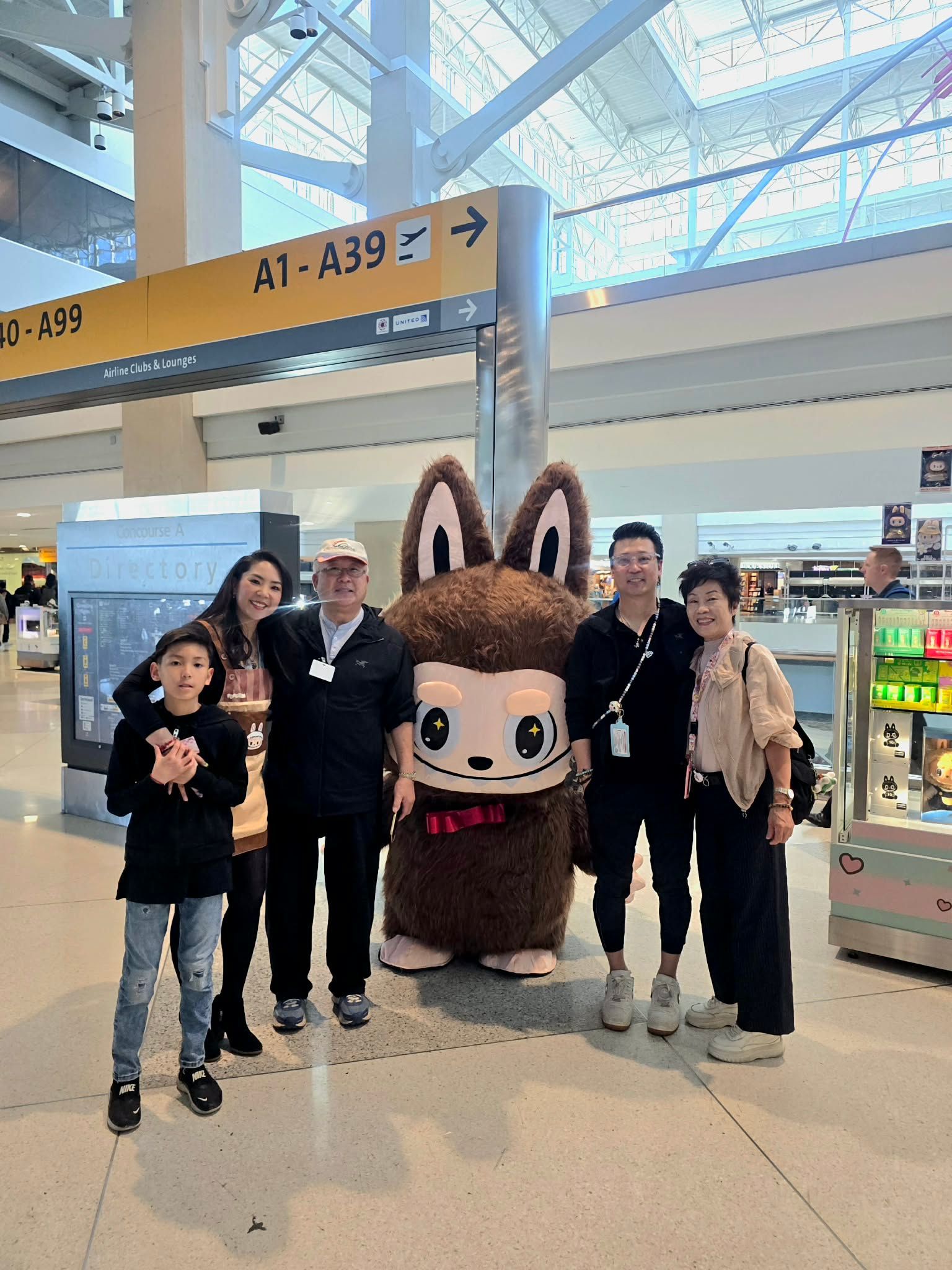 A group of six people posing for a photo with a large brown mascot character in an airport terminal near gate signs.
