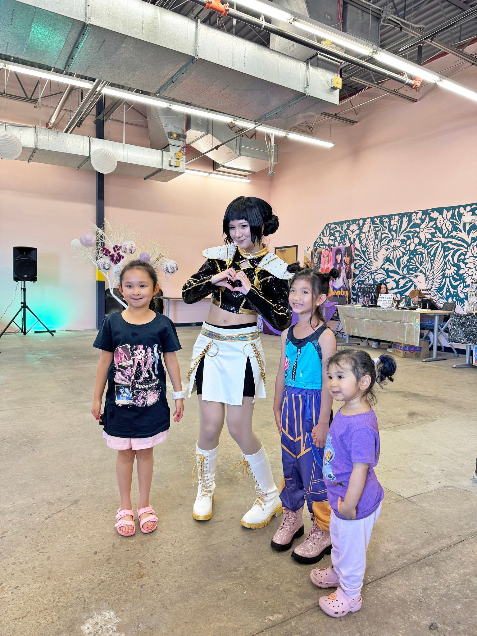 A costumed person poses with three young children. Indoor setting, pink wall, black speaker, and light blue.
